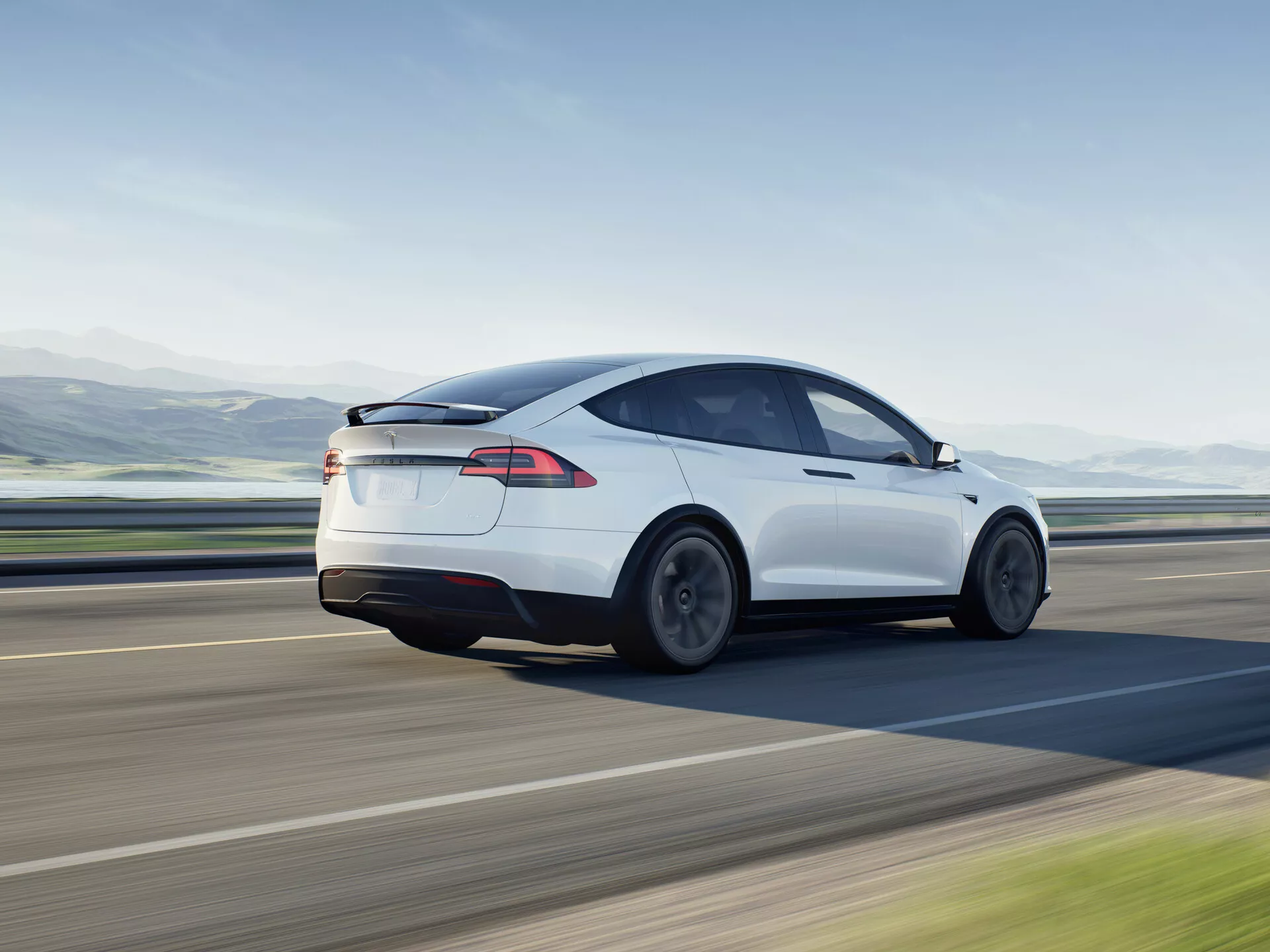 Image dynamique d'une berline électrique blanche Tesla Model X 2021 en mouvement, vue depuis un angle trois-quarts arrière. La photographie capture le côté gauche du véhicule depuis une perspective légèrement surélevée, mettant en évidence les roues arrière, les feux arrière et le contour de l'extrémité arrière, soulignant son design aérodynamique avec une ligne de toit inclinée et des courbes lisses. Le design indique une turbulence d'air minimisée et un faible coefficient de traînée, soutenu par des roues conçues pour l'efficacité aérodynamique. La lunette arrière se fond dans un toit en verre noir, suggérant un toit panoramique, symbole de la technologie automobile moderne. Les élégants feux arrière à LED sont une caractéristique distinctive de la technologie d'éclairage contemporaine. L'absence de tuyau d'échappement indique la nature entièrement électrique du véhicule. Idéal pour des catégories telles que véhicules, véhicules électriques, photographie de mouvement, conception automobile, technologie moderne et véhicules de luxe.