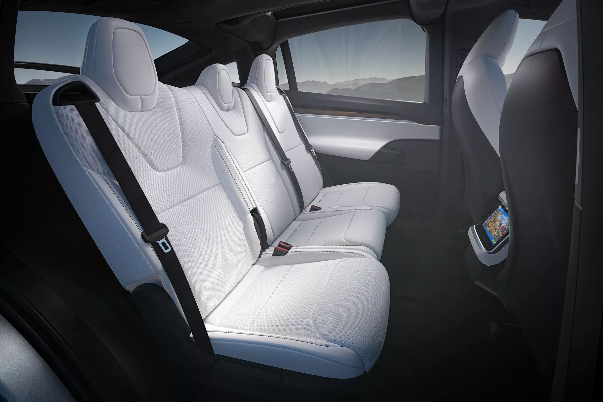 Vue intérieure d'une Tesla Model X 2021 (1ère génération-facelift), montrant le compartiment passagers arrière depuis la porte arrière côté conducteur. Sièges en cuir blanc avec appuie-tête et rembourrage profilé pouvant accueillir trois passagers. Un écran numérique au dos du siège passager avant suggère un système de divertissement à l'arrière. L'habitacle spacieux, possiblement doté d'un toit panoramique, utilise la lumière naturelle pour accentuer les panneaux de porte sombres avec des grilles de haut-parleurs intégrées.