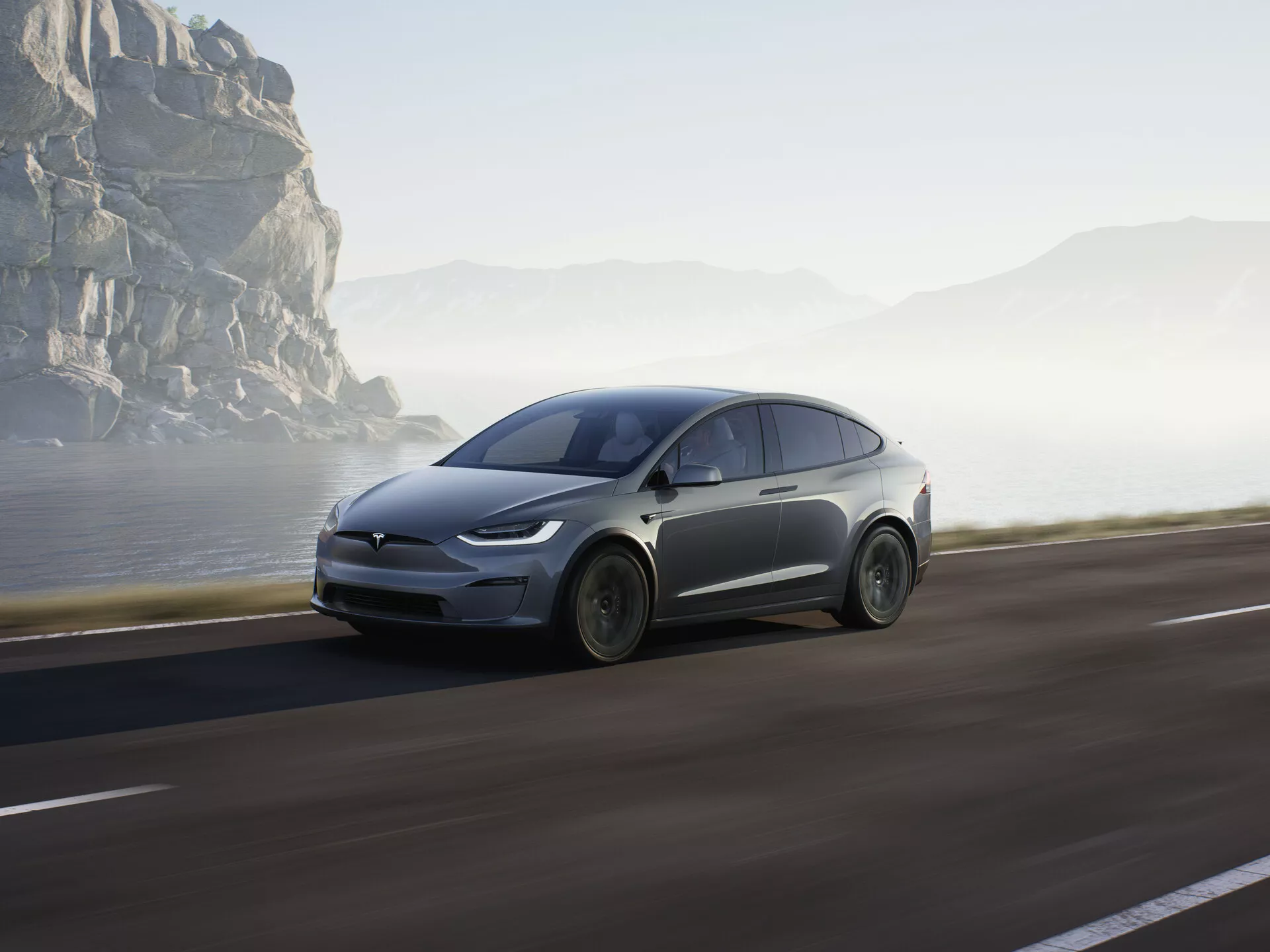L'image montre un SUV électrique moderne, spécifiquement une Tesla Model X 2021 (facelift de première génération), roulant le long d'une route côtière. Il est capturé en vue de profil bas, mettant en évidence le quart avant avec un accent sur la roue avant, le capot, le pare-brise et une partie de la porte du conducteur. Le véhicule occupe une grande partie de la moitié inférieure du cadre, soulignant sa proéminence. La voiture est en mouvement avec un léger flou de mouvement autour des roues et de l'arrière-plan, créant une sensation de vitesse et de dynamisme. La lumière douce suggère tôt le matin ou en fin d'après-midi, ajoutant une atmosphère tranquille à l'environnement côtier pittoresque et aventureux. Des éléments de design aérodynamique et des fonctionnalités avancées telles que les phares à LED peuvent être inférés, indiquant sa technologie avancée comme le freinage régénératif et peut-être des capacités de conduite autonome. Positionnée sur le côté droit de la route, la voiture avance vers une destination ouverte, les rochers côtiers et le brouillard à l'arrière-plan renforçant son attrait futuriste et son cadre naturel.