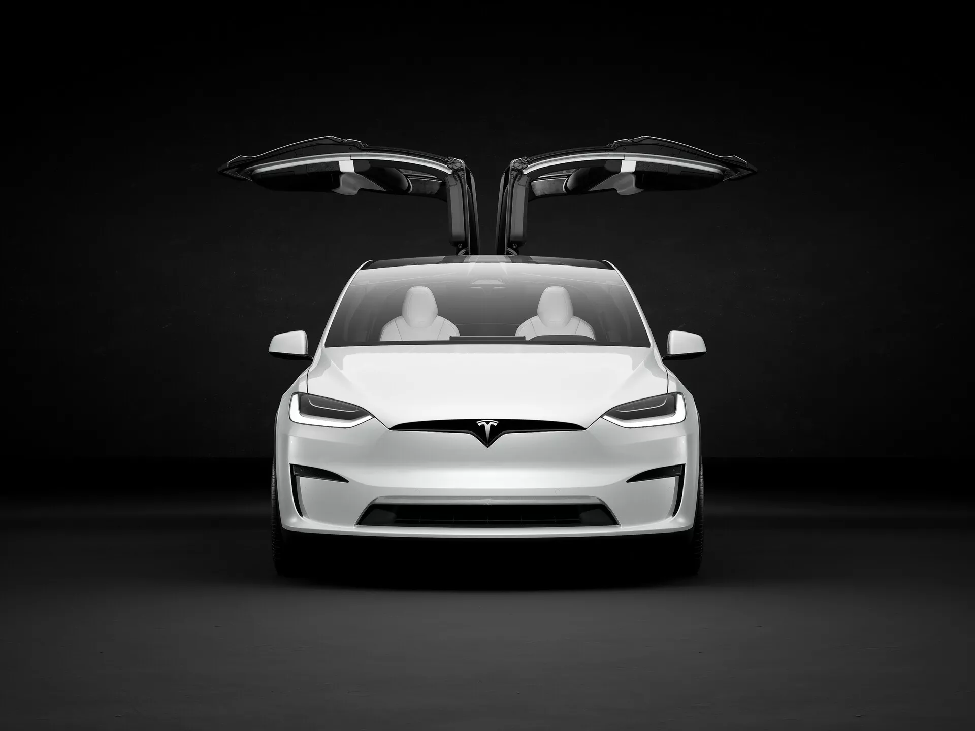 L'image présente un véhicule électrique Tesla Model X 2021 (première génération, lifting) dans un environnement de studio avec un fond sombre qui contraste avec la couleur claire de la voiture. Vue légèrement sous un angle bas, la partie avant de la voiture est mise en valeur, montrant le capot, le pare-chocs et la zone de la calandre, bien que cette dernière ne soit pas traditionnelle puisqu'il s'agit d'un véhicule électrique. La prise d'air inférieure est également visible. Les deux portes avant sont ouvertes, révélant les portes Falcon-Wing qui s'ouvrent vers le haut, une caractéristique distinctive des modèles haut de gamme. L'intérieur montre deux rangées de sièges de couleur claire, suggérant un design minimaliste mais luxueux. Les phares à LED anguleux et tranchants ajoutent une touche agressive, et le logo Tesla est placé de manière proéminente au-dessus de la prise d'air et sous le 'frunk'. La voiture laisse entrevoir des fonctionnalités avancées telles que la conduite autonome, le groupe motopropulseur électrique, les systèmes de freinage régénératif, la connectivité et les systèmes de sécurité avancés, visant un public qui valorise l'innovation, la performance et la conscience environnementale.