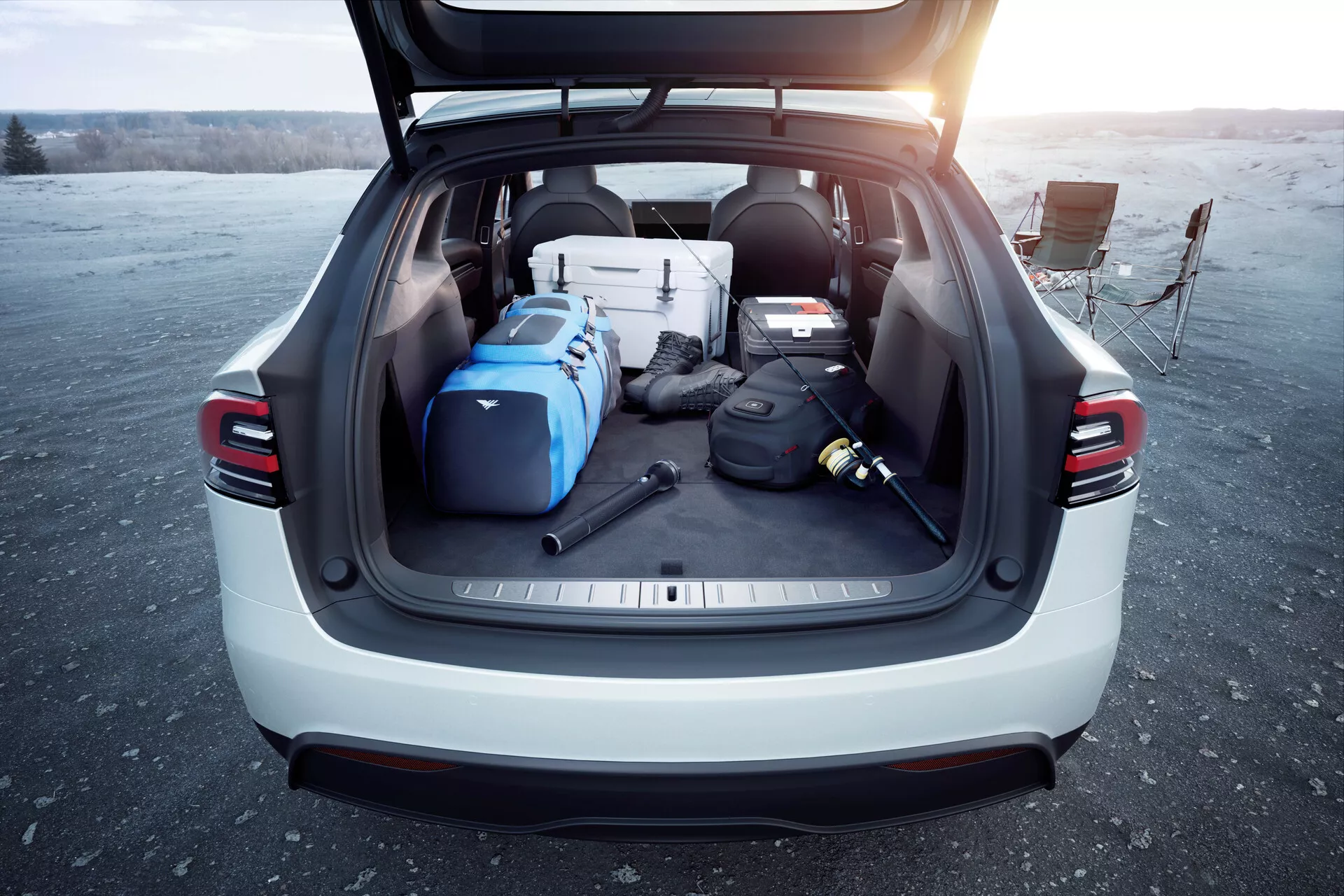 Vue arrière d'un SUV Tesla Model X 2021 avec le coffre ouvert, montrant son espace de chargement spacieux rempli de divers objets sous la lumière du jour, peut-être au lever ou au coucher du soleil. L'image met en avant l'utilité et les caractéristiques modernes du véhicule telles que le hayon automatique, les feux arrière à LED, la couleur extérieure claire et le couvre-charge, prise dans un espace ouvert.