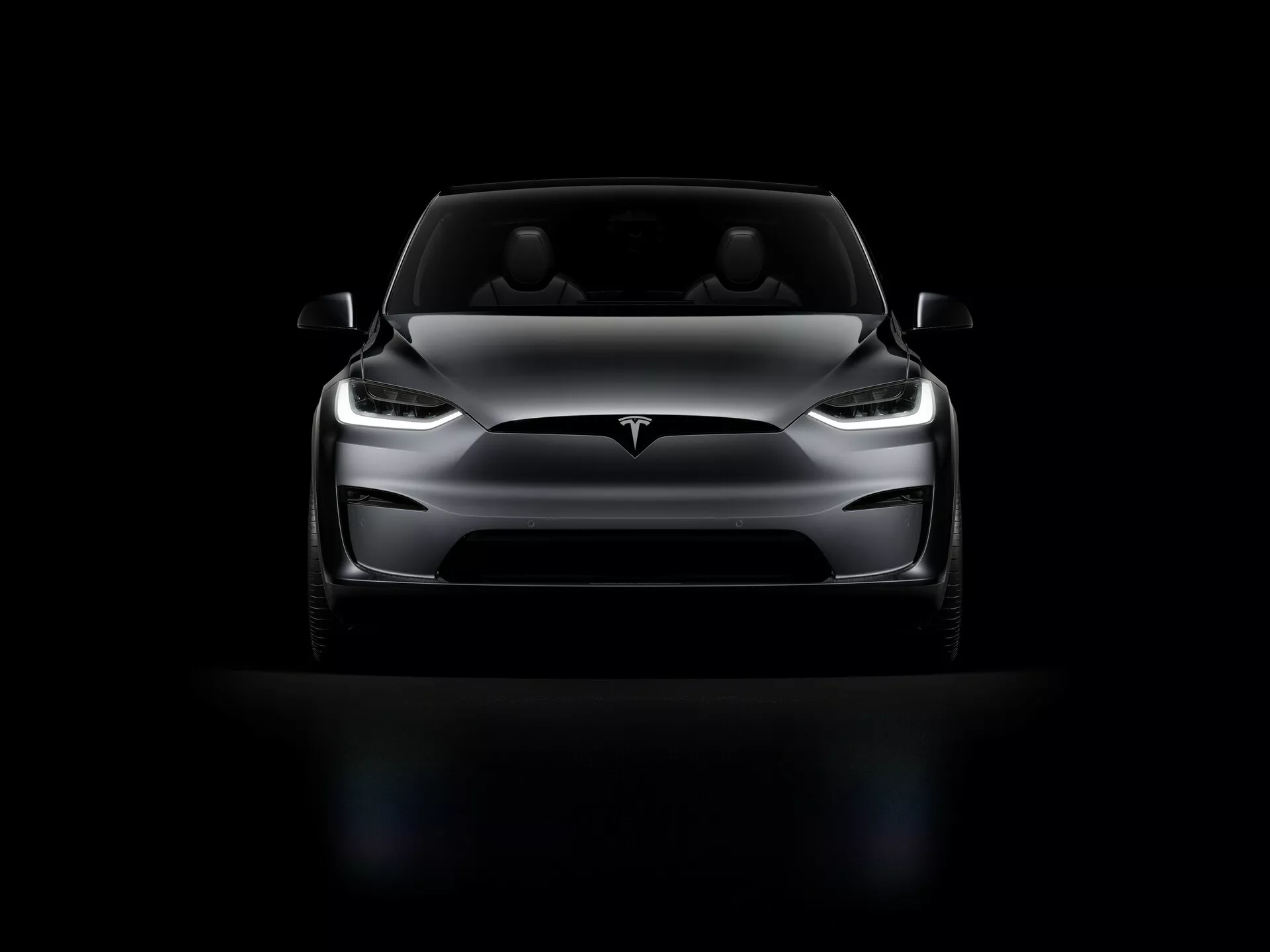 Tesla Model X 2021 (1ère génération facelift) photographiée de face avec un angle bas. L'image met en avant la face avant de la voiture, les phares LED, le design élégant du capot, l'avant lisse sans calandre, le pare-chocs avant minimaliste et les rétroviseurs latéraux aérodynamiques. L'éclairage dramatique met en lumière les caractéristiques clés, tandis que la voiture est placée contre un fond noir, accentuant sa posture imposante.