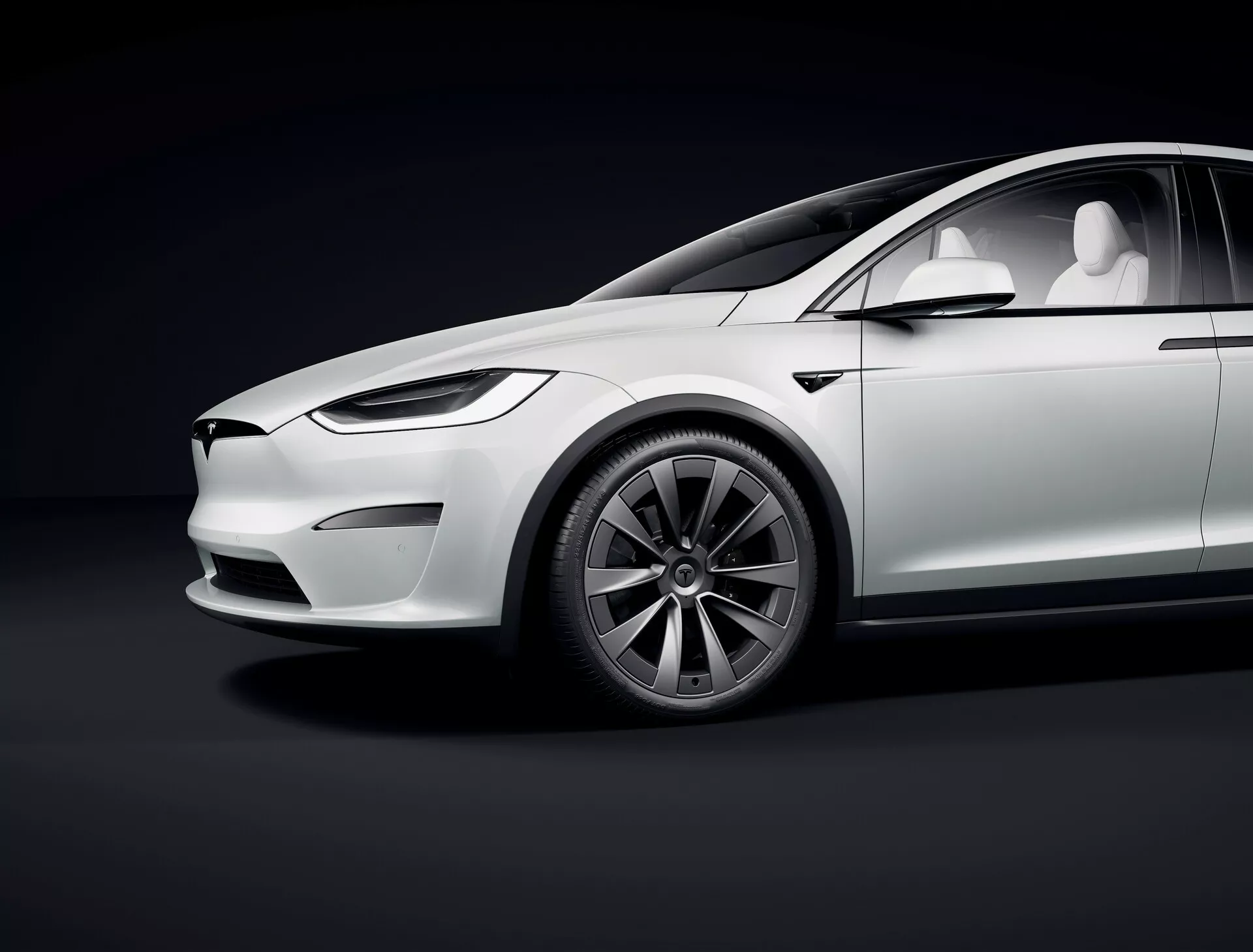 Photo en studio montrant le profil latéral de la Tesla Model X 2021 première génération remise à niveau, sous un angle bas, mettant en avant des caractéristiques de luxe électrique telles que le design épuré, absence de tuyaux d'échappement, avant minimaliste avec phares LED, poignées de portes rétractables, roues à motifs aérodynamiques multi-rayons, grands pneus à profil bas, peinture blanche brillante avec des reflets doux, toit panoramique et rétroviseurs basés sur caméra. Les courbes et lignes du véhicule sont accentuées par l'éclairage du studio sur un fond dégradé foncé.