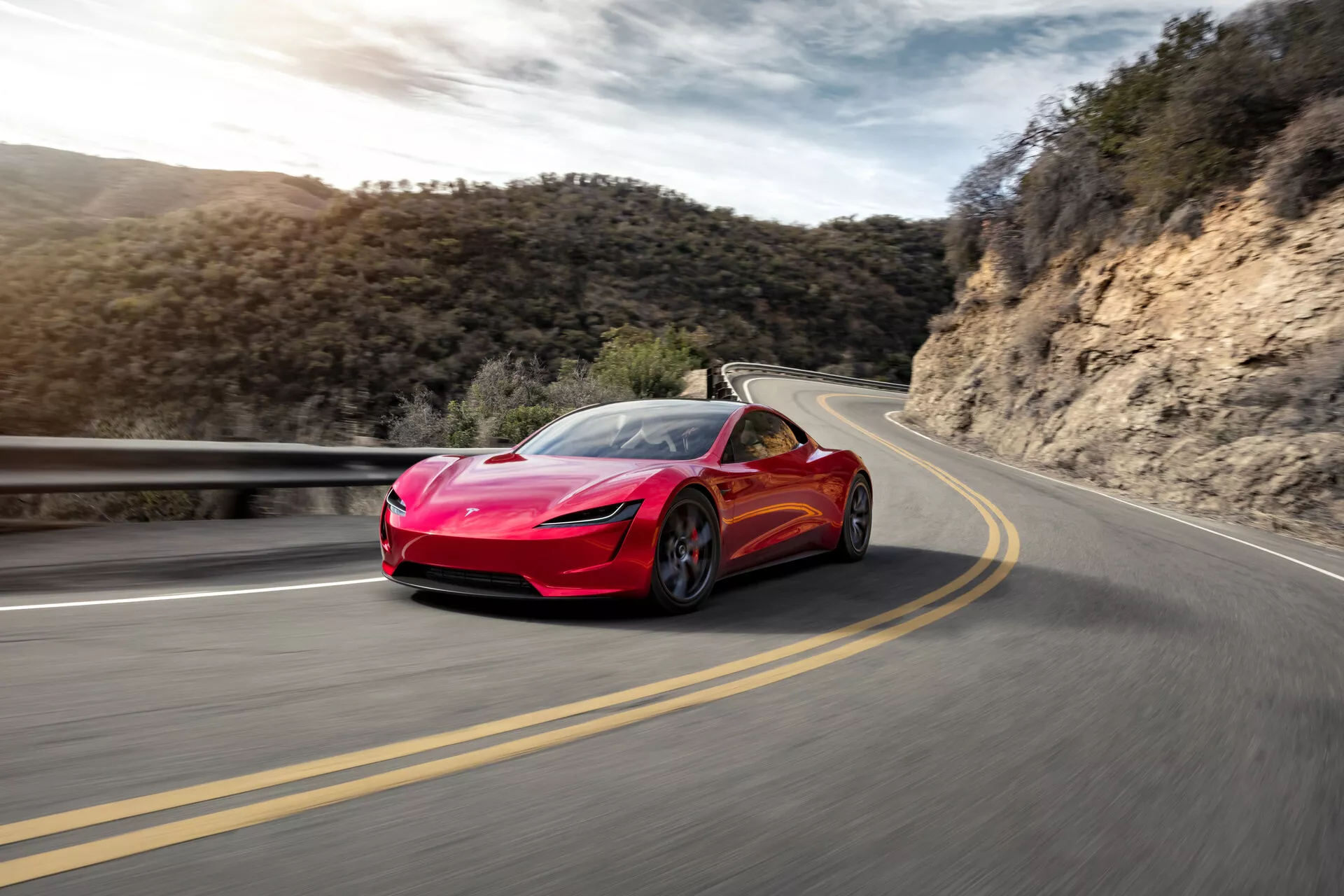 Image d'une voiture de sport Tesla Roadster 2020 (2ème génération) rouge vif en mouvement sur une route sinueuse. La vue de trois quarts avant capture des phares élégants, une grille aérodynamique, de grandes prises d'air avant et des lignes sculptées sur le capot. L'apparence imposante de la voiture est accentuée par un angle de prise de vue bas. La route floue et l'arrière-plan suggèrent une haute vitesse, avec la voiture positionnée sur le côté droit de la route, sur fond de ciel clair et de végétation.