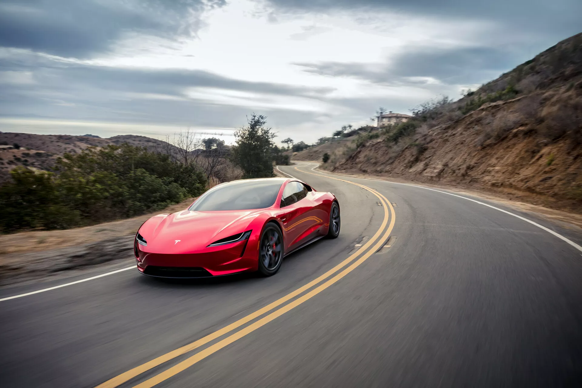 Photo dynamique d'une voiture sport Tesla Roadster 2020 (deuxième génération) rouge capturée en mouvement sur une route sinueuse montrant ses lignes aérodynamiques, ses phares élégants, ses rétroviseurs, ses grandes roues et ses prises d'air, contre un arrière-plan de lumière diffuse indiquant une conduite à grande vitesse dans un environnement rural.