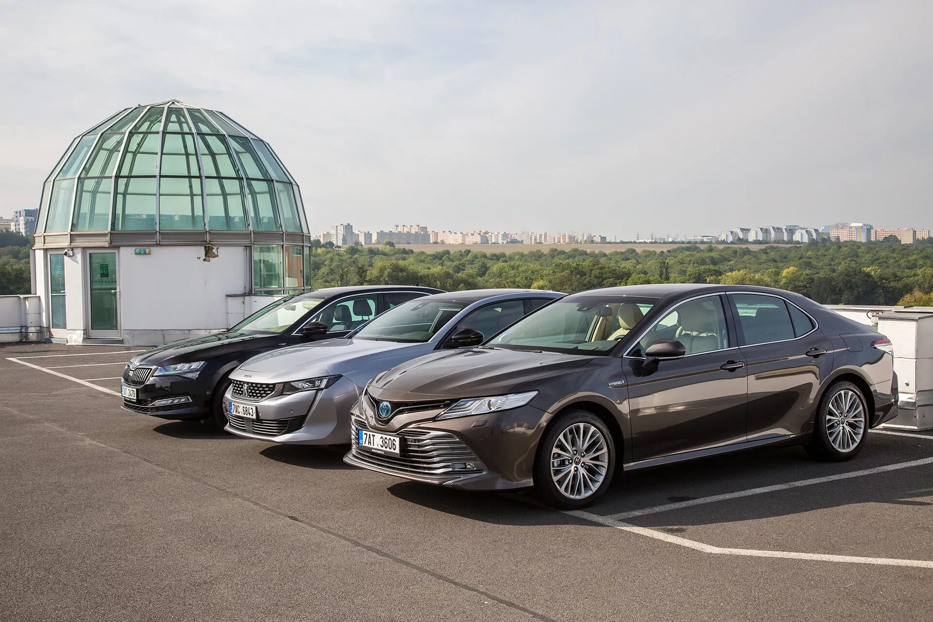 Limousine Toyota Camry 2019 garée sur un pont de toit avec ciel clair et dôme de verre en arrière-plan, présentant un design moderne avec une grille avant dynamique, des phares LED pointus, un capot sculpté et des jantes en alliage de luxe