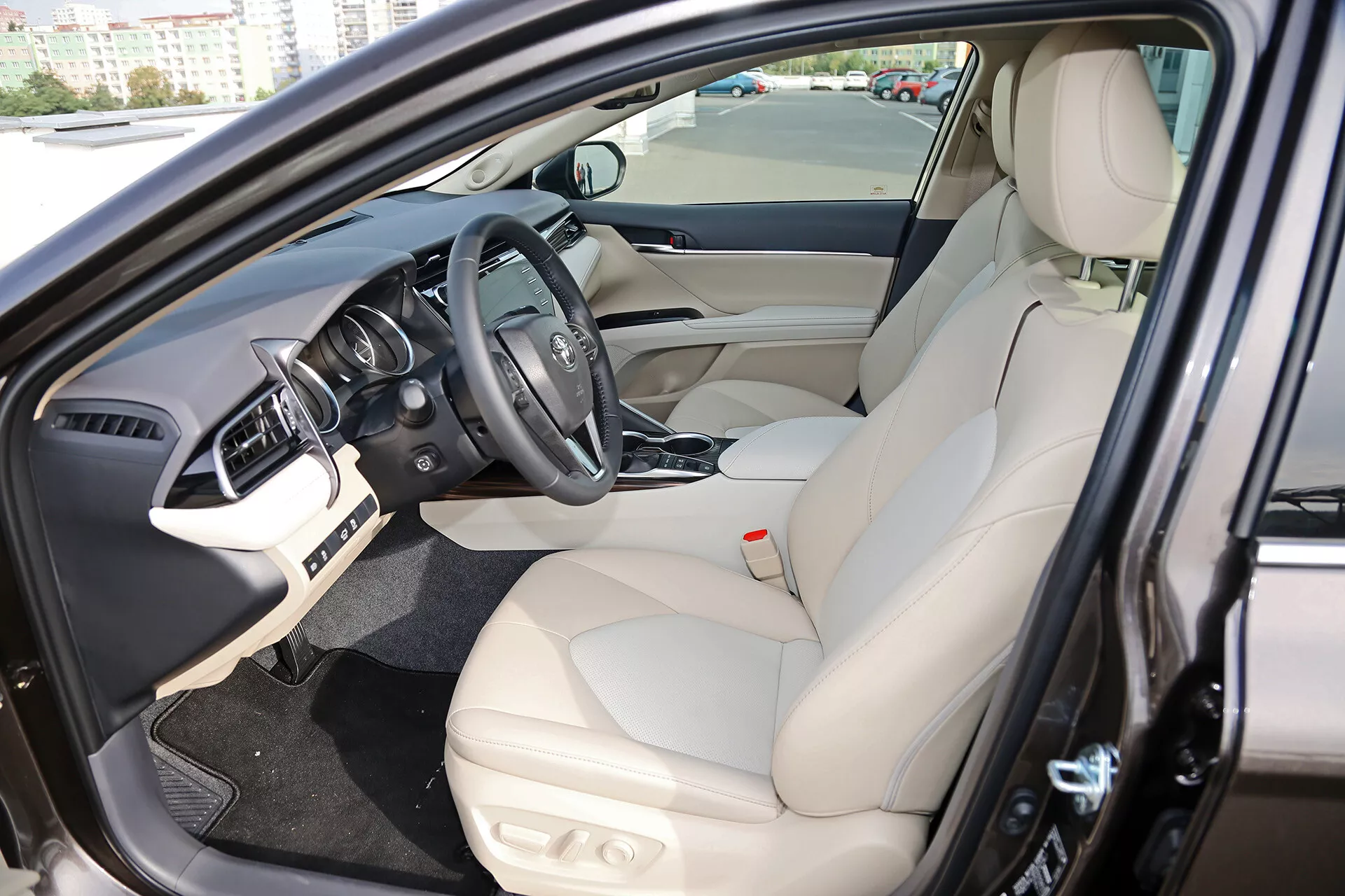 Intérieur de la Limousine Toyota Camry 2001 montrant la zone du conducteur depuis un angle frontal oblique avec la porte ouverte, mettant en évidence le siège du conducteur en cuir avec des commandes électroniques, le volant central avec des commandes montées et le logo du fabricant, le tableau de bord avec écran d'infodivertissement et le panneau intérieur de la porte avec des commandes pour les vitres électriques.