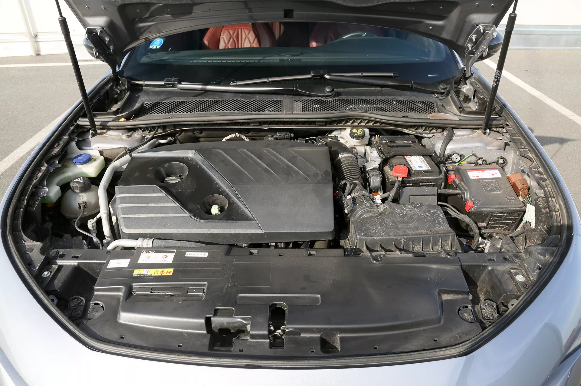 Vue de face du compartiment moteur d'une Toyota Camry 2001 moderne, montrant le couvercle du moteur avec logo, batterie de 12 volts, câblage et tuyaux complexes, réservoir de refroidissement et boîte à fusibles dans une configuration détaillée indiquant une technologie moteur avancée et maintenance.