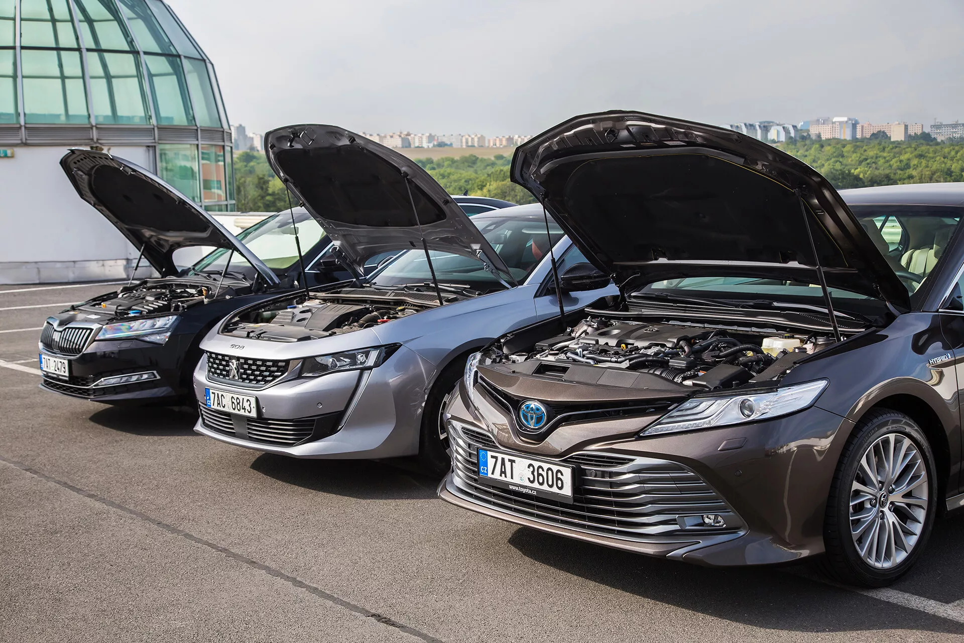 Alignement de trois voitures diverses avec les capots ouverts montrant les moteurs, incluant une Škoda argentée, un SUV Peugeot gris et une Toyota Camry gris foncé, vue en angle contre un arrière-plan urbain avec une structure de façade en verre et un horizon de ville, mettant en lumière la technologie et les caractéristiques de conception des véhicules.