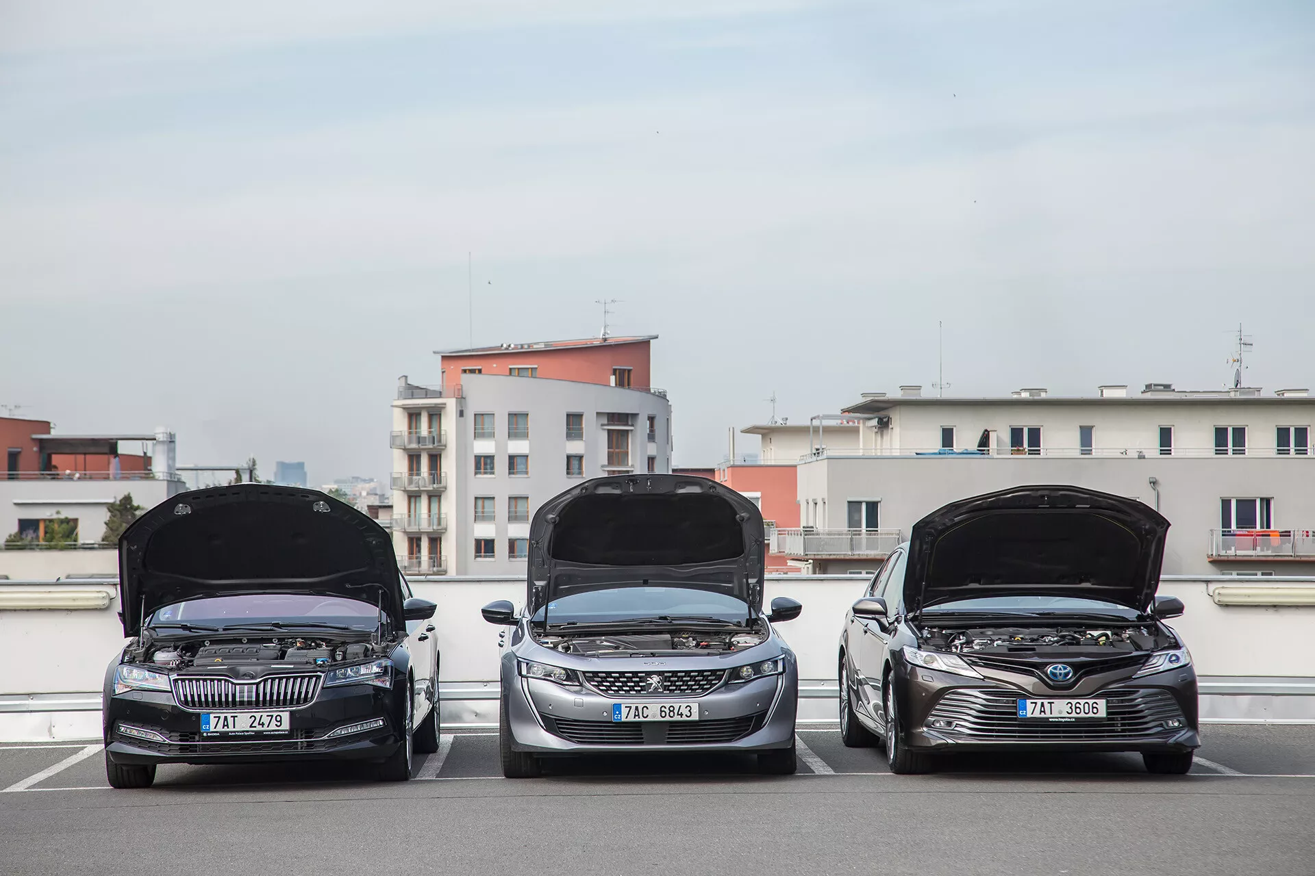 Trois limousines Toyota Camry de 2001 garées côte à côte sur un parking en rooftop, vue de face avec capots ouverts révélant les compartiments moteur, lumière du jour mettant en valeur les caractéristiques technologiques et le design sur un fond de ville