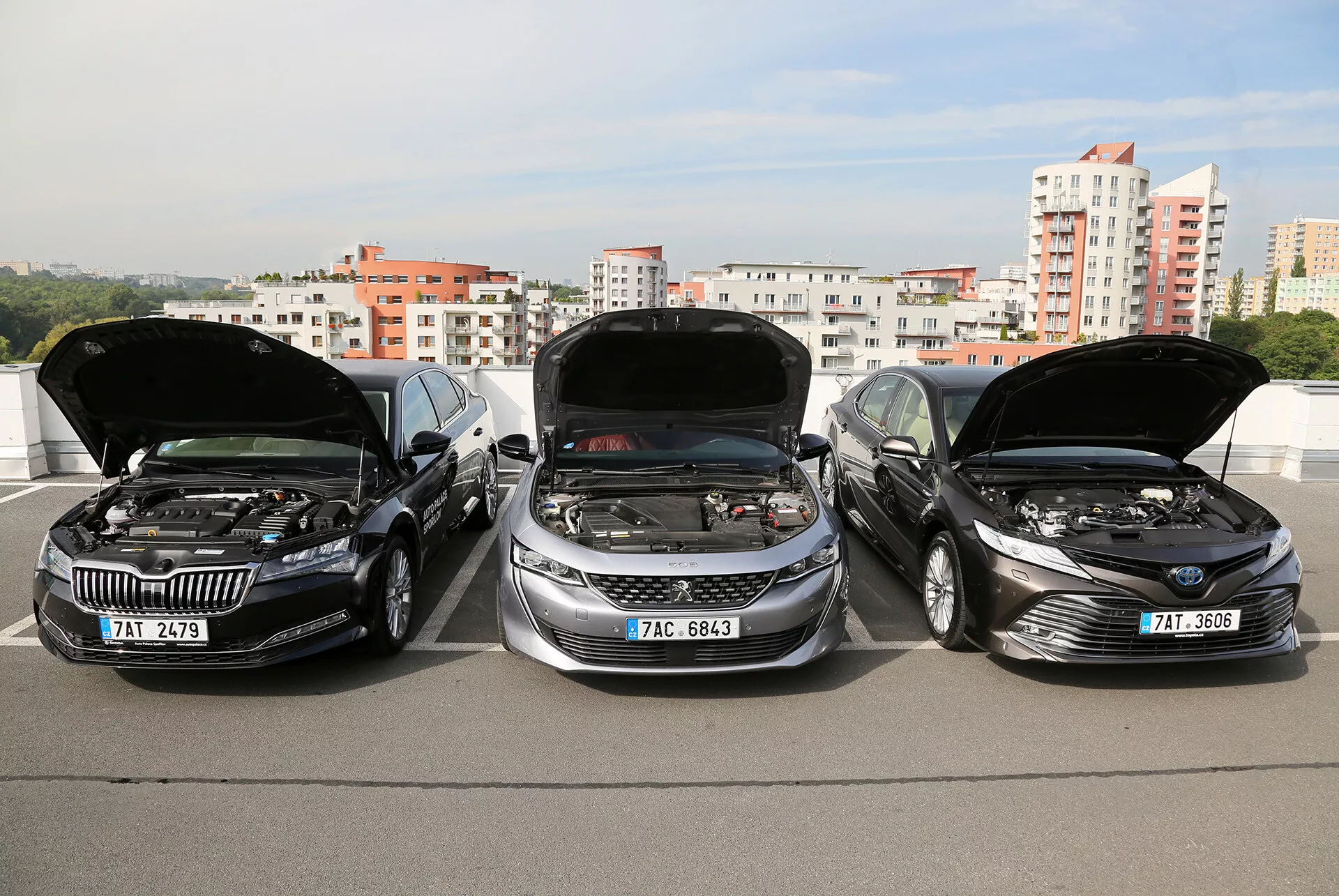 Modèles de limousine Toyota Camry 2019 avec les capots ouverts montrant les moteurs, garés côte à côte sur une structure surélevée, avec un horizon de ville en arrière-plan, mettant en avant le design épuré, moderne des véhicules et la technologie avancée du moteur.