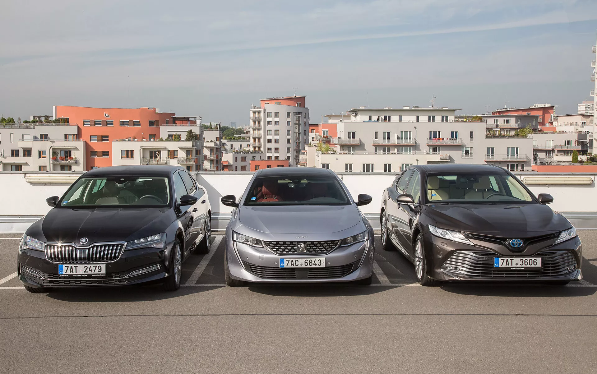 Comparaison côte à côte de berlines Skoda, Peugeot et Toyota dans une vue de trois quarts avant sur un parking en rooftop, mettant en évidence des caractéristiques de design moderne telles que l'éclairage LED, les formes aérodynamiques et les accents chromés