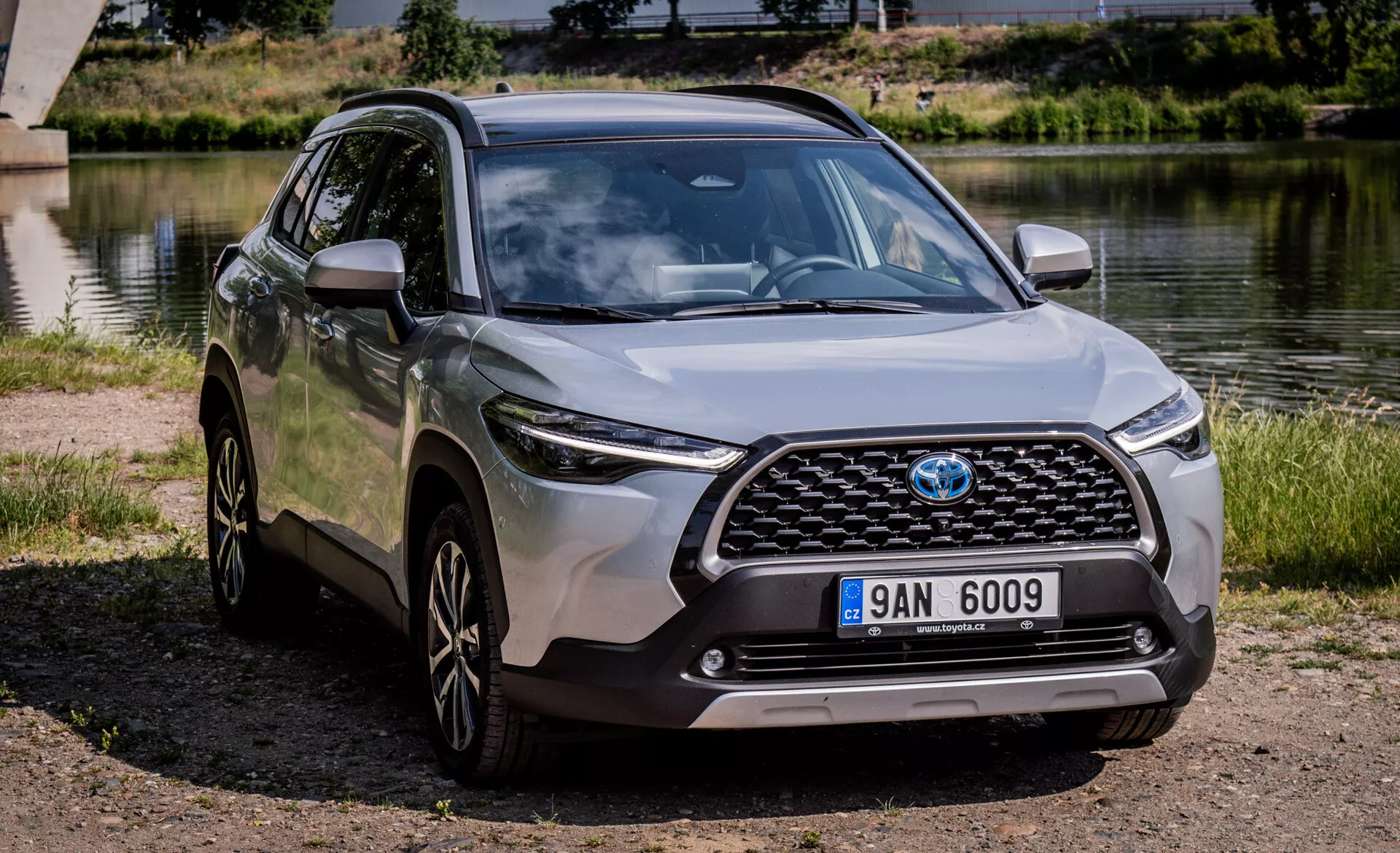 SUV crossover moderne Toyota Corolla Cross 2022 stationné en extérieur près d'un plan d'eau offrant une vue claire de sa grille avant, capot, phares LED, pare-chocs avant, et profil latéral incluant jantes en alliage et rétroviseurs latéraux couleur carrosserie