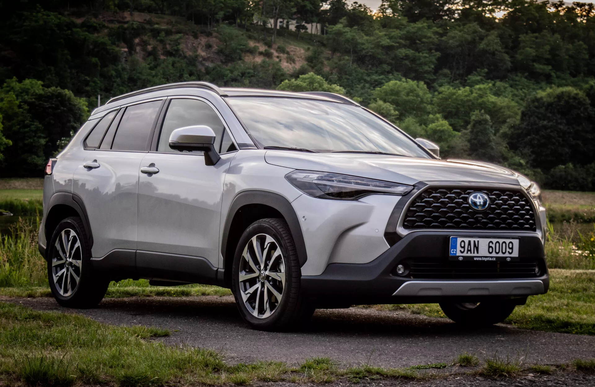 Toyota Corolla Cross 2022 en finition métallique brillante montrant son design moderne de SUV avec grille avant détaillée, emblème, phares LED angulaires, pare-choc avant aérodynamique, capot profilé, profil latéral avec roues en alliage à plusieurs branches et barres de toit, prise depuis un angle bas pour souligner sa hauteur et sa posture durant le jour dans un cadre suburbain naturel.