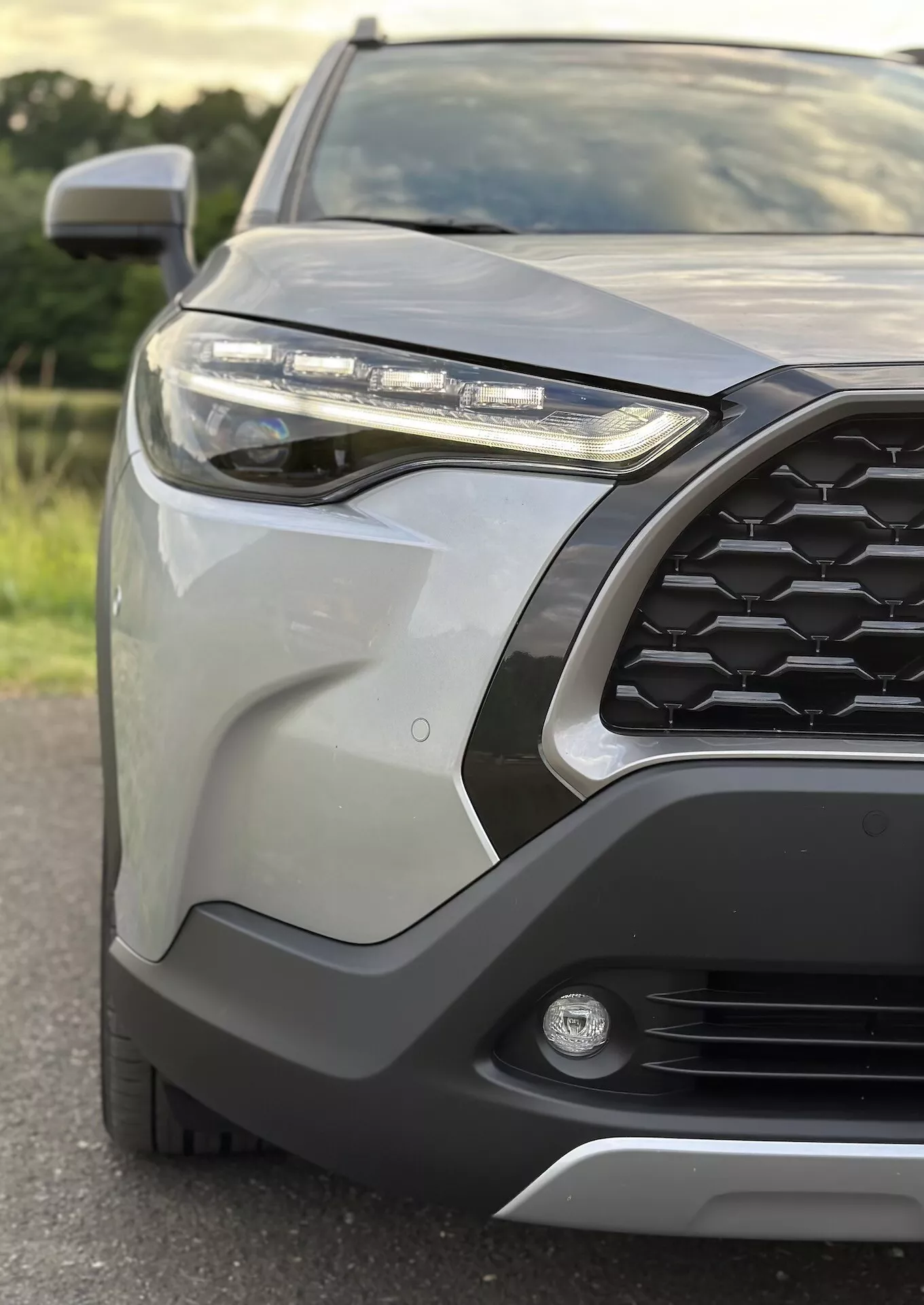 Gros plan sur le quart avant-gauche du Toyota Corolla Cross 2022 mettant en évidence la grille hexagonale, le phare LED, le feu de brouillard, le pare-chocs avant avec revêtement protecteur, les panneaux de carrosserie élégants, le rétroviseur latéral avec clignotant intégré et la finition métallique