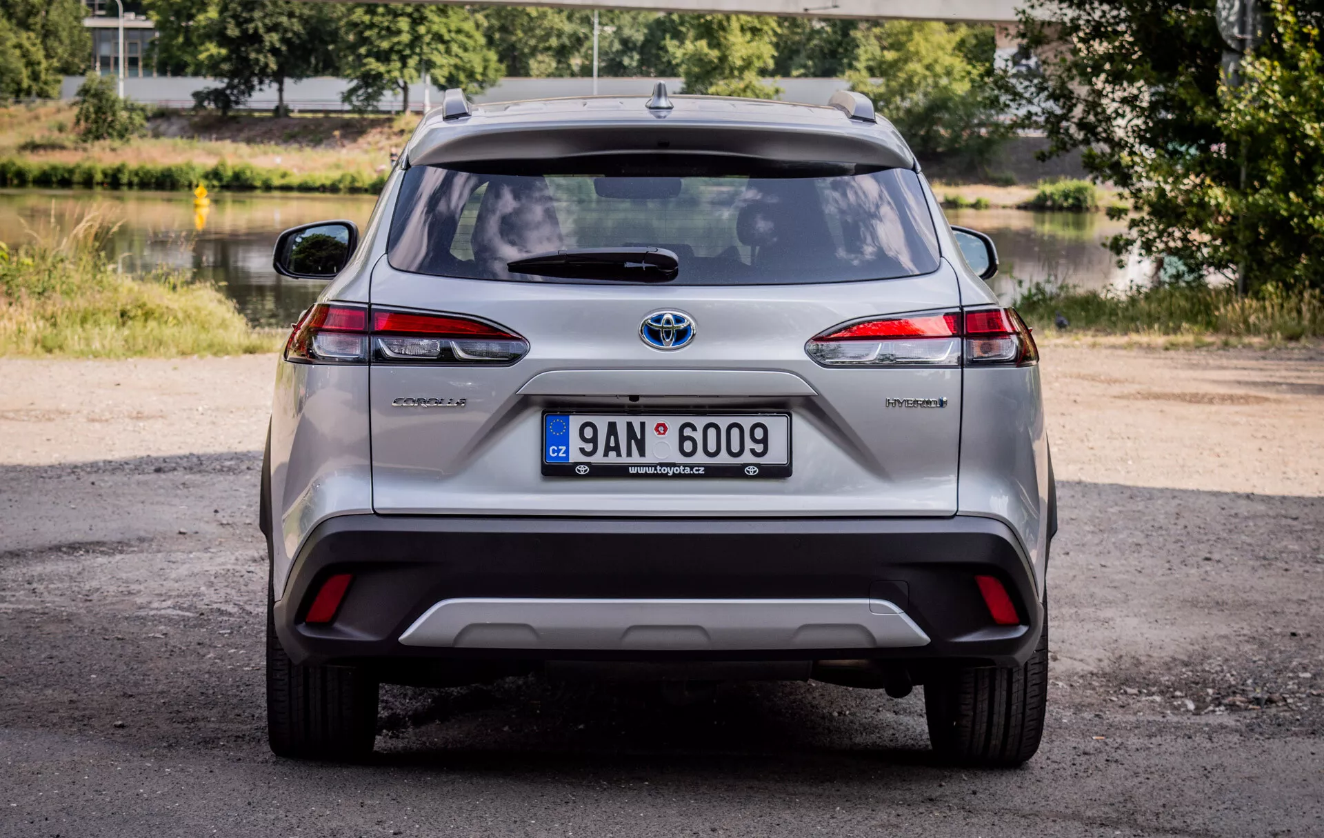 Vue arrière de la Toyota Corolla Cross 2022, présentant des feux arrière LED, un badge hybride et un emblème Toyota chromé, avec un éclairage naturel de jour et un arrière-plan pittoresque de verdure et d'eau