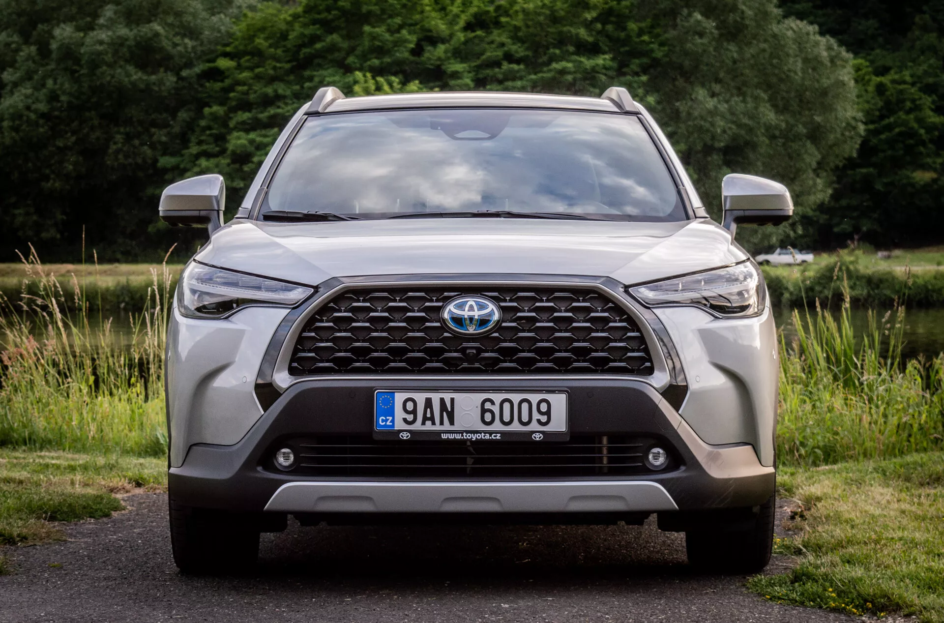 Vue de face d'une Toyota Corolla Cross 2022, mettant en évidence sa grande grille avec emblème, phares modernes à LED, feux de brouillard, position de la plaque d'immatriculation, capteurs de stationnement intégrés ou caméra frontale, contours aérodynamiques du capot, design robuste du pare-chocs inférieur et prises d'air, stationnée sur l'asphalte avec un arrière-plan naturel