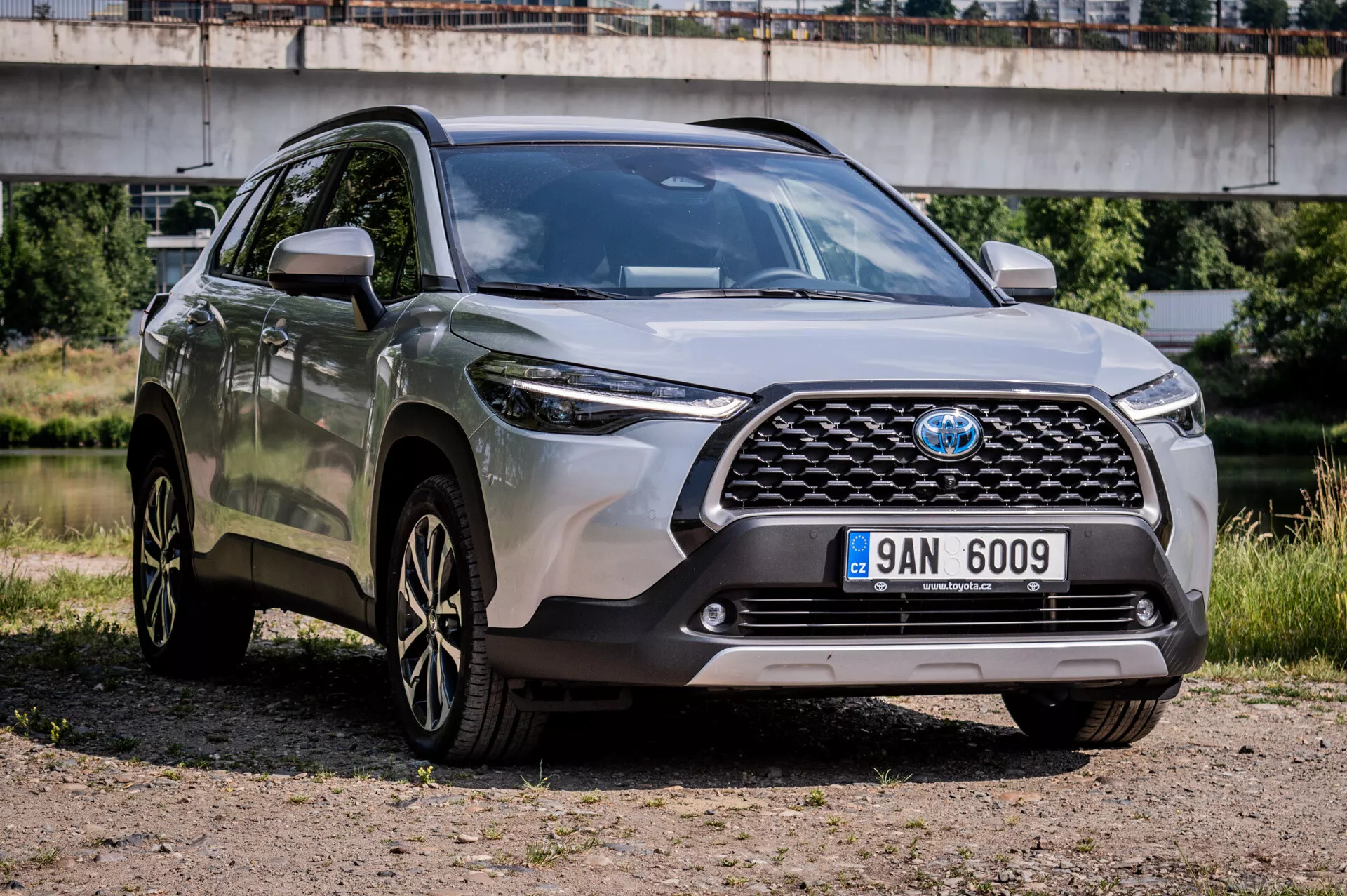 Toyota Corolla Cross 2022 garée sur gravier montrant son design robuste, ses jantes en alliage sportif à multi-rayons, ses phares LED avancés et sa garde au sol élevée dans une vue avant aux trois quarts mettant en évidence la grille avant, le pare-chocs et les caractéristiques du côté conducteur avec un pont en arrière-plan semi-urbain