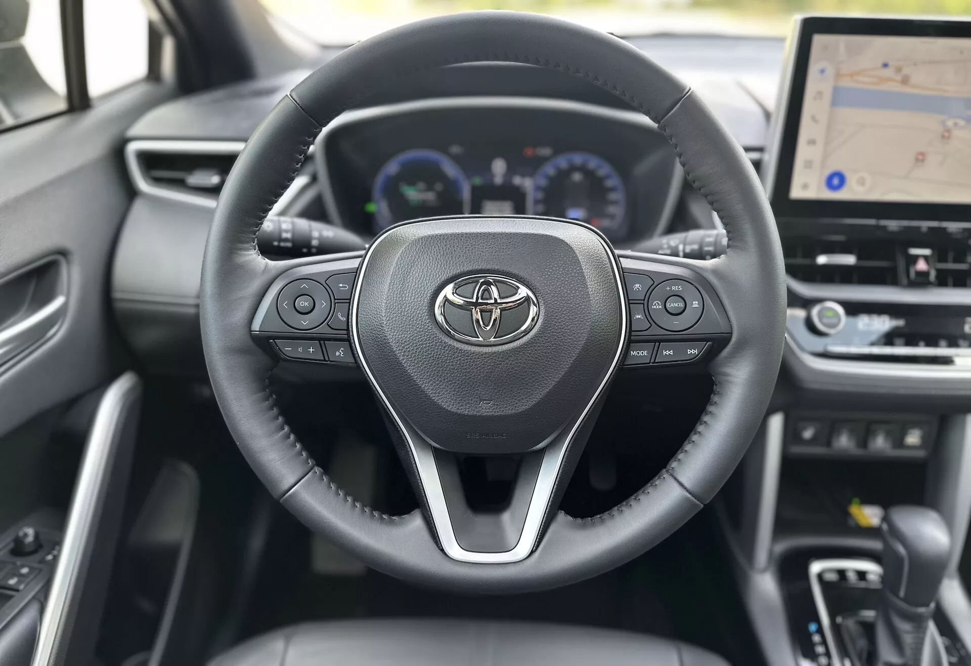Intérieur de la Toyota Corolla Cross 2022 montrant le volant avec le logo Toyota, les commandes intégrées pour l'audio, les appels et le régulateur de vitesse, derrière lequel un tableau de bord numérique avec compteur de vitesse, compte-tours et affichage multifonctions est partiellement visible, y compris un affichage central plus grand pour la navigation GPS, le tout sur fond de design contemporain en matériaux sombres.