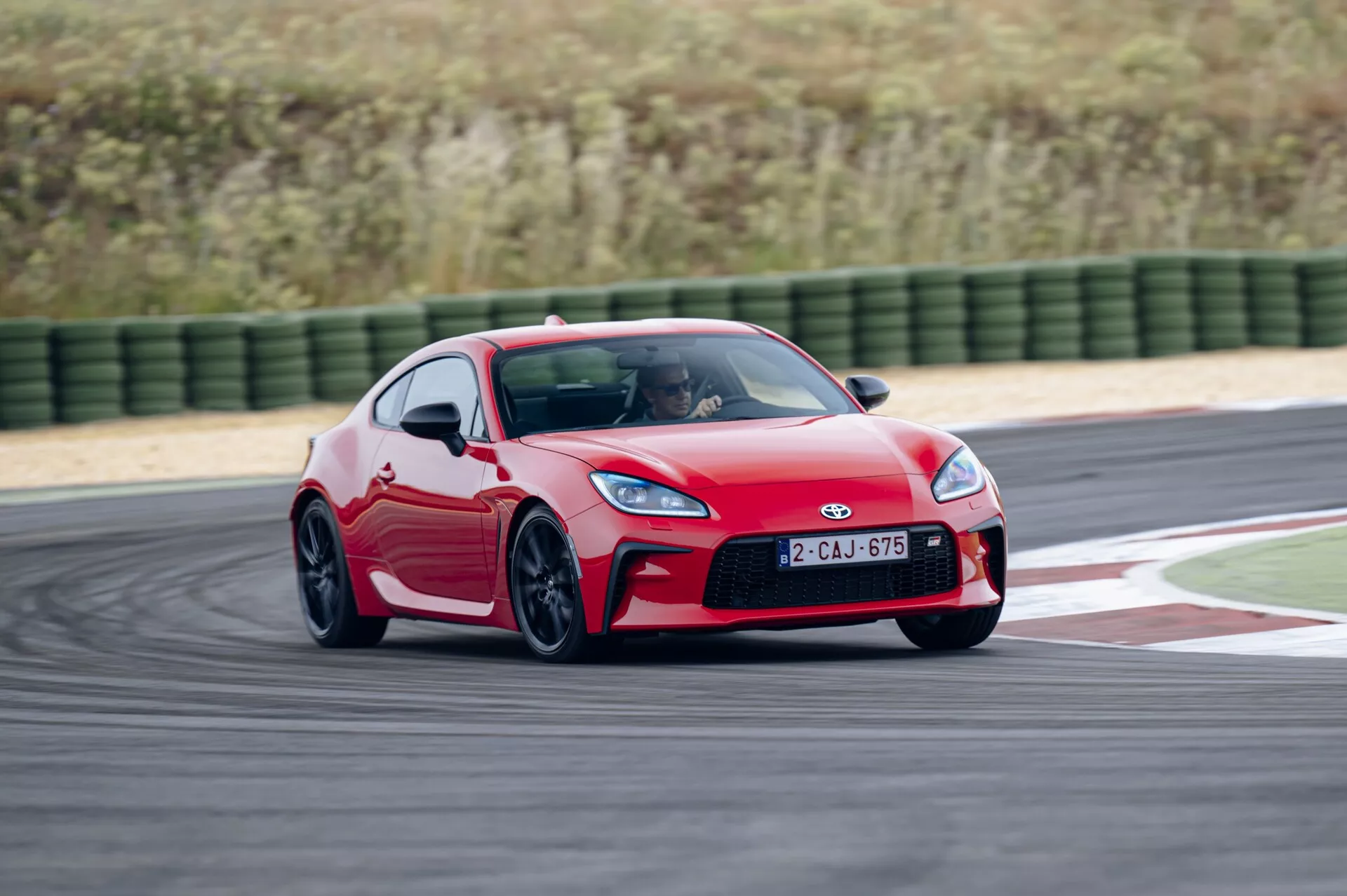 Une prise de vue dynamique d'une Toyota GT86 2022 rouge capturée sous un angle avant trois quarts en mouvement sur une piste de course, montrant le design aérodynamique, les phares à LED, les pneus haute performance et les lignes aérodynamiques de la voiture le long du côté, suggérant une technologie automobile avancée et une maniabilité routière supérieure.
