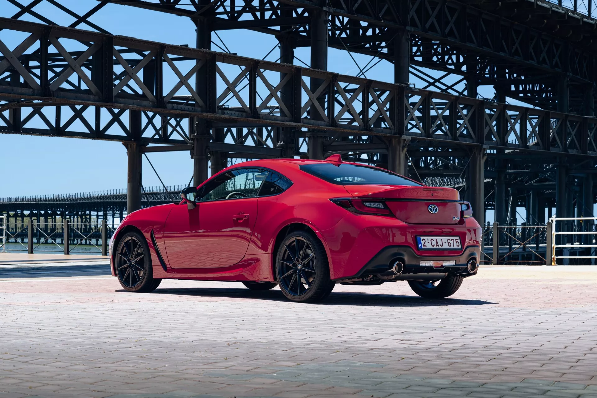 La Toyota GT86 2022, vue de trois quarts arrière, montrant le côté droit. Les ailes arrière musclées, les feux arrière LED modernes, les tuyaux d'échappement intégrés dans le pare-chocs inférieur et les jantes multi-bras en alliage performantes avec des étriers de frein visibles sont mis en avant. La peinture métallisée et les pneus profil bas soulignent le caractère sportif et haute performance de la voiture, capturé sur un arrière-plan industriel.