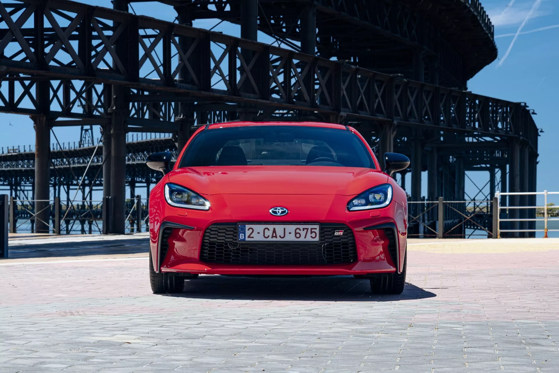 L'image montre une Toyota GT86 2022 rouge capturée sous un angle frontal bas, mettant en avant la large et agressive calandre du véhicule, les phares élégants probablement LED et le pare-chocs avant avec prises ou évents, suggérant une meilleure aérodynamique ou refroidissement. Le capot présente des lignes profilées menant à l'emblème, soulignant son design aérodynamique et orienté vers la performance. La voiture est positionnée sur une zone pavée avec un arrière-plan industriel, suggérant un environnement urbain ou semi-urbain.