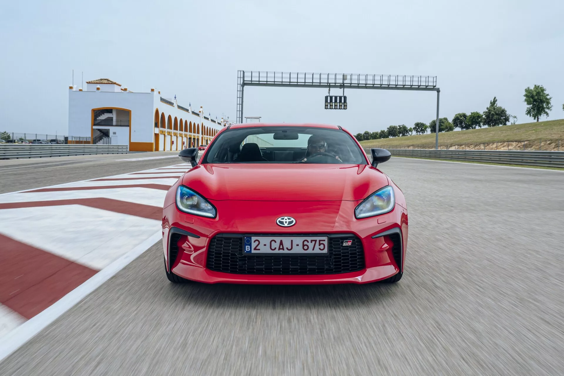 Vue de face d'une voiture de sport Toyota GT86 rouge de 2022 sur un circuit, avec une grande calandre centrale, des phares au design agressif et des prises d'air visibles sur le pare-chocs avec une structure de départ/arrivée en arrière-plan.