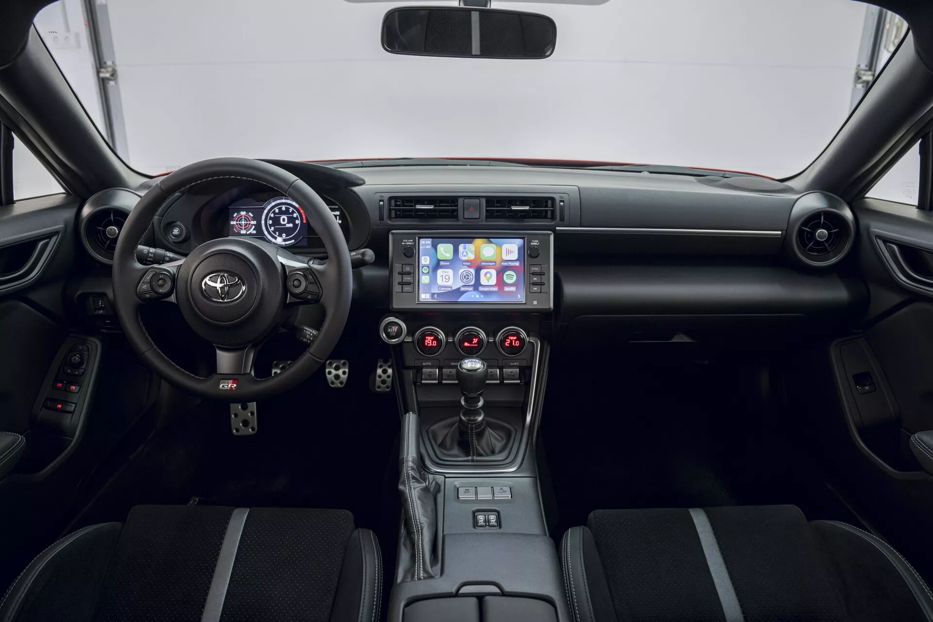 L'intérieur de la Toyota GT86 2022 est présenté depuis la perspective du siège arrière, offrant une vue frontale symétrique du tableau de bord. L'appareil photo, positionné approximativement à la hauteur du tableau de bord, met en évidence le volant avec les commandes intégrées et le badge Toyota proéminent. Derrière le volant se trouve un affichage numérique qui affiche probablement la vitesse et les informations du conducteur. La console centrale comprend un écran tactile d'infodivertissement de 6 à 8 pouces affichant une interface de menu colorée avec la navigation, l'audio et les paramètres du véhicule. Plus bas se trouvent des bouches d'aération et des boutons rouges pour le contrôle du climat. Le levier de vitesses automatique est accompagné de boutons supplémentaires et de deux porte-gobelets. Les sièges avant en tissu bicolore et le motif de motif ajoutent à l'apparence sophistiquée, avec des commandes sur les portes et des accents argentés sur le tableau de bord, soulignant l'intérieur moderne et orienté vers le confort du véhicule.