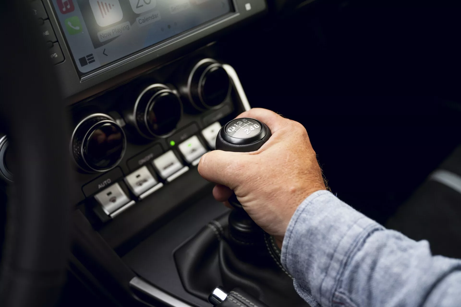 Vue rapprochée depuis le siège passager avant de la console centrale de la Toyota GT86 2022, se concentrant sur la zone du levier de vitesses. L'image capture la main d'une personne conduisant, apparemment masculine, saisissant le levier de vitesses manuel en position neutre. La console mélange des matériaux plastiques noirs et en cuir avec des accents métalliques ou chromés, ainsi que des boutons indiquant des technologies modernes telles que les capteurs de stationnement et les systèmes stop-start.