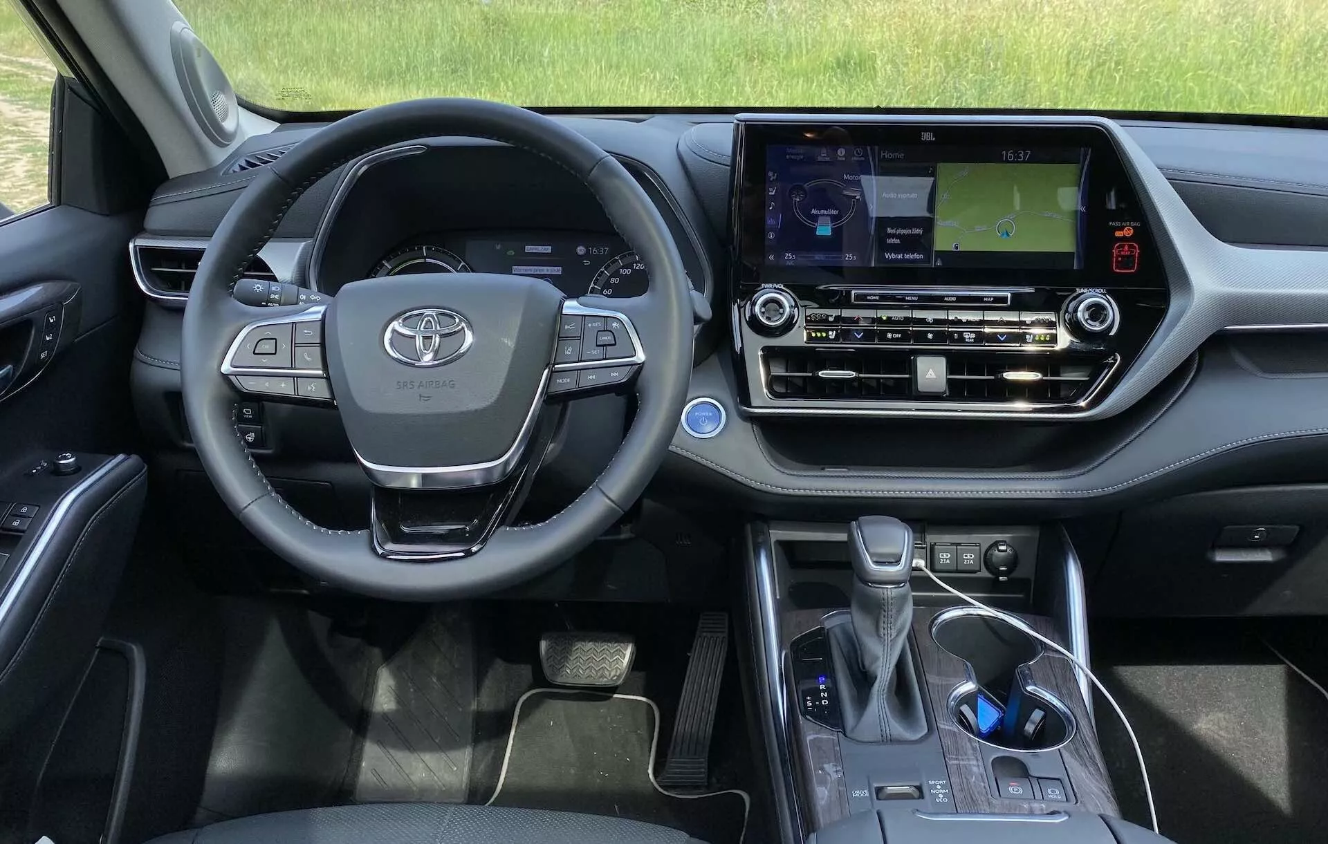 L'intérieur de la Toyota Highlander 2.5 Hybrid Business Edition 2021 du point de vue du conducteur. L'image montre le volant avec l'emblème Toyota, des boutons multifonctionnels, le tableau de bord partiellement visible et la console centrale avec un système d'infodivertissement à écran tactile affichant une carte de navigation. Les commandes de climat, un compartiment de rangement, le levier de changement de vitesse électronique et le frein de stationnement électronique sont également visibles, reflétant le design moderne et les caractéristiques technologiques du véhicule.