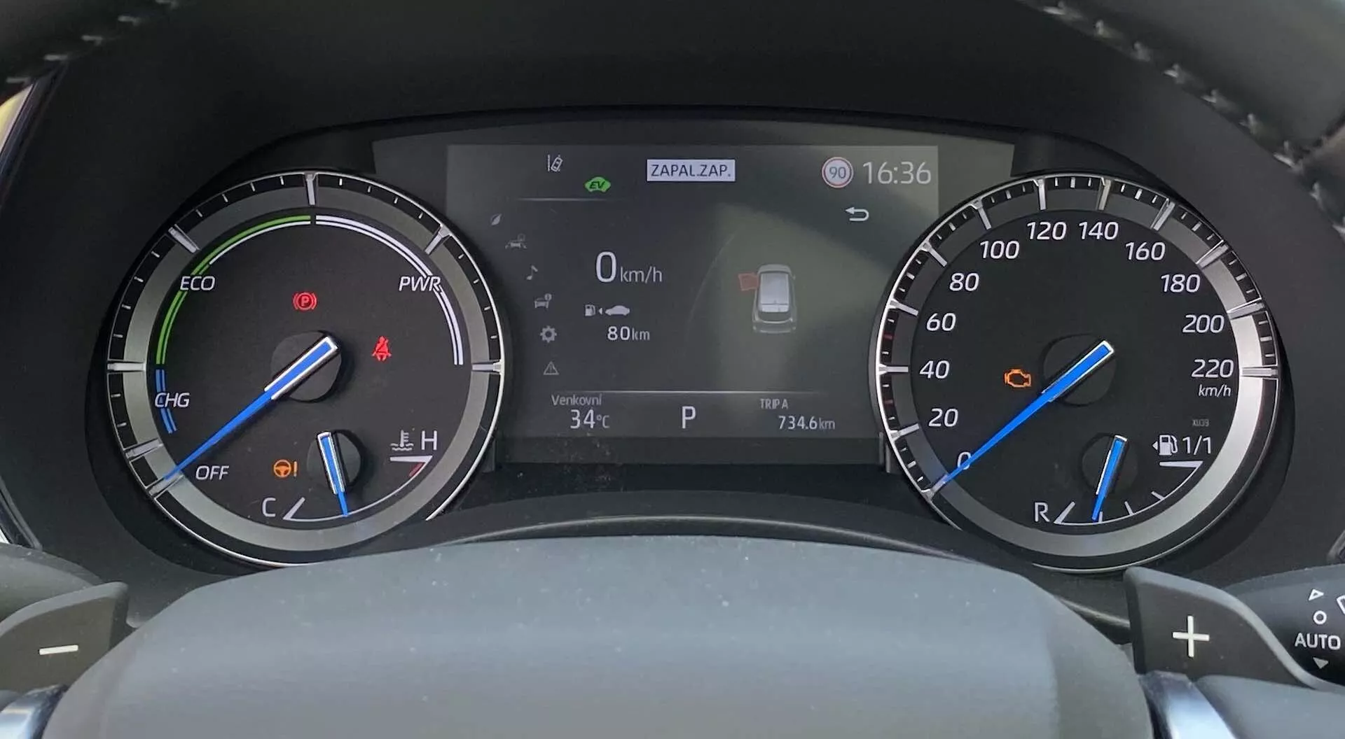 Tableau de bord hybride digital-analogique du Toyota Highlander 2021, vu du siège du conducteur, avec un compteur de régime avec indicateur de mode ECO à gauche, un compteur de vitesse avec indicateur de niveau de carburant à droite et un écran central digital affichant la vitesse (0 km/h), la température extérieure (34°C), l'horloge (16:36) et une icône du système d'assistance à la conduite.