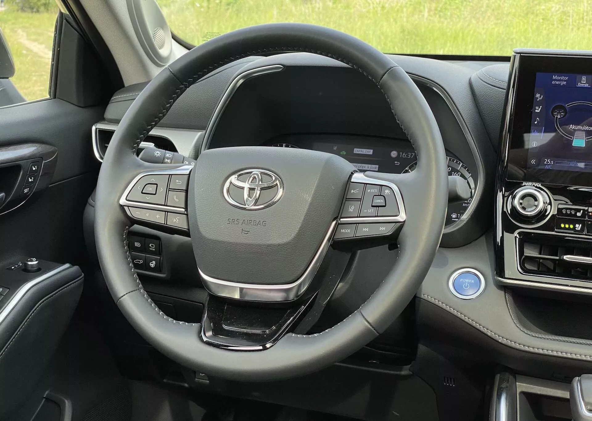 Vue intérieure du Toyota Highlander 2.5 Hybrid Business Edition 2021 depuis une perspective en première personne, focalisée sur le volant multifonction avec commandes audio, connectivité téléphonique et régulateur de vitesse, avec le logo Toyota clairement visible au centre. Le tableau de bord, la console centrale avec système d'infotainment tactile, le levier de vitesse, le panneau de contrôle de la porte et la ventilation sont visibles, mettant en avant des matériaux de haute qualité et des fonctionnalités avancées.