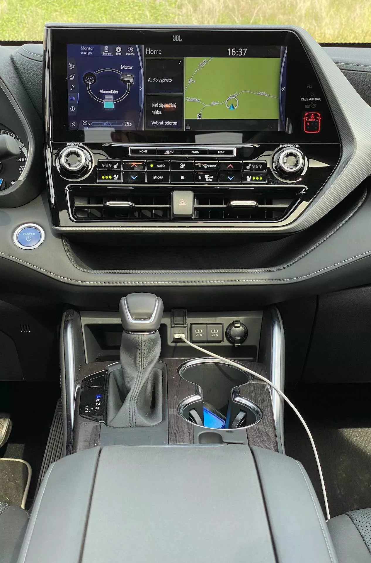 Vue intérieure de la Toyota Highlander 2.5 Hybrid Business Edition 2021 depuis le siège arrière, avec un système d'infodivertissement moderne à écran tactile affichant une horloge et une carte de navigation, des boutons physiques pour le contrôle du climat, des évents d'air, un levier de vitesses automatique avec garnissage en cuir et accents argentés, un bouton de commande, un accoudoir central avec rangement ouvert, deux porte-gobelets, un câble de charge USB et un design contemporain bicolore avec des accents métalliques.