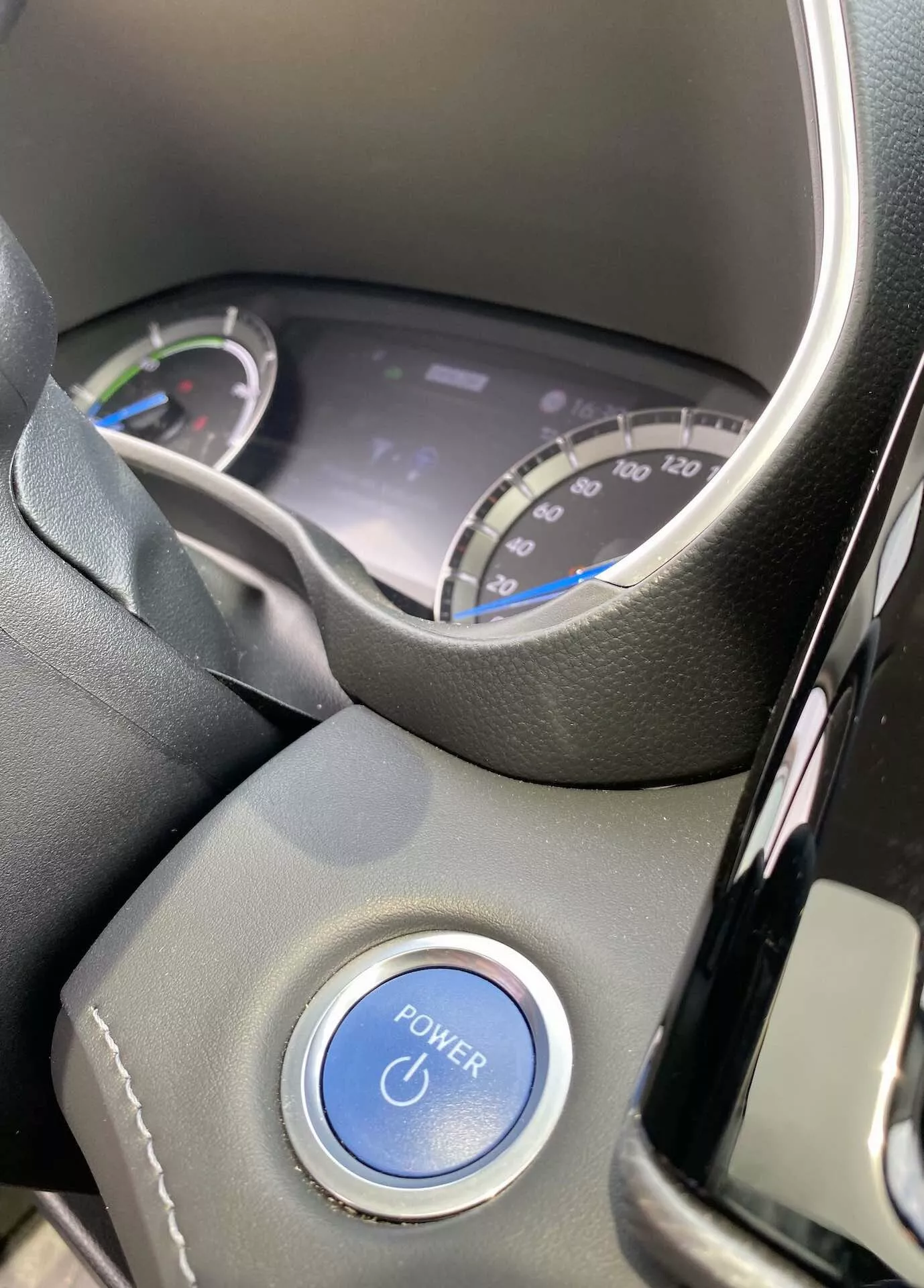 Vue intérieure du Toyota Highlander 2021 2.5 Hybrid Business Edition axée sur la perspective du conducteur. L'image montre une partie du volant et le tableau de bord numérique à travers la section supérieure du volant. Le tableau comprend un compteur de vitesse marqué jusqu'à 220 km/h et un cadran de consommation d'énergie, suggérant la nature hybride du véhicule. Un bouton bleu 'POWER' est visible sur la colonne de direction, soulignant le système de démarrage sans clé. L'intérieur présente des plastiques noirs modernes et un volant en cuir ou semblable, avec des conditions de lumière naturelle accentuant l'état neuf ou bien entretenu de la voiture.