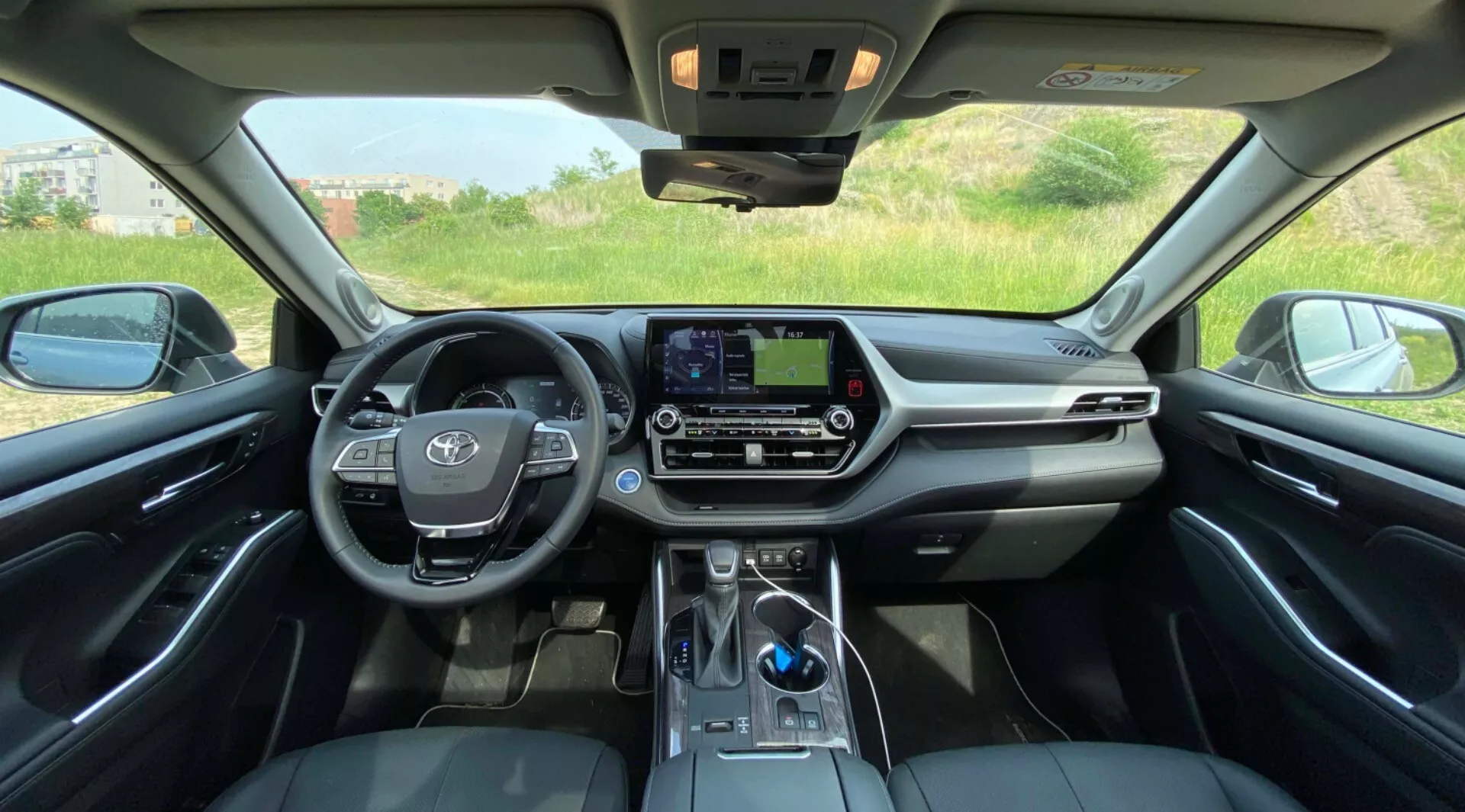 Vue intérieure du Toyota Highlander 2021 Hybrid Business Edition depuis le siège arrière, montrant une perspective symétrique du tableau de bord avant. Le volant, avec l'emblème du véhicule et des commandes audio intégrées, est bien visible sur la gauche. Le centre du tableau de bord dispose d'un écran d'infodivertissement tactile sans boutons physiques, accompagné de boutons et de boutons de commande climatique en dessous. Le sélecteur de vitesses, un levier électronique, est visible dans la partie inférieure centrale, près d'un éventuel espace de charge pour smartphones. L'intérieur luxueux se distingue par une combinaison de sièges en cuir, de matériaux doux au toucher sur le tableau de bord et des accents chromés. Le toit ouvrant permet à la lumière naturelle de pénétrer, avec des panneaux de commande près du rétroviseur. À l'extérieur, on aperçoit un paysage naturel à travers les fenêtres avant.