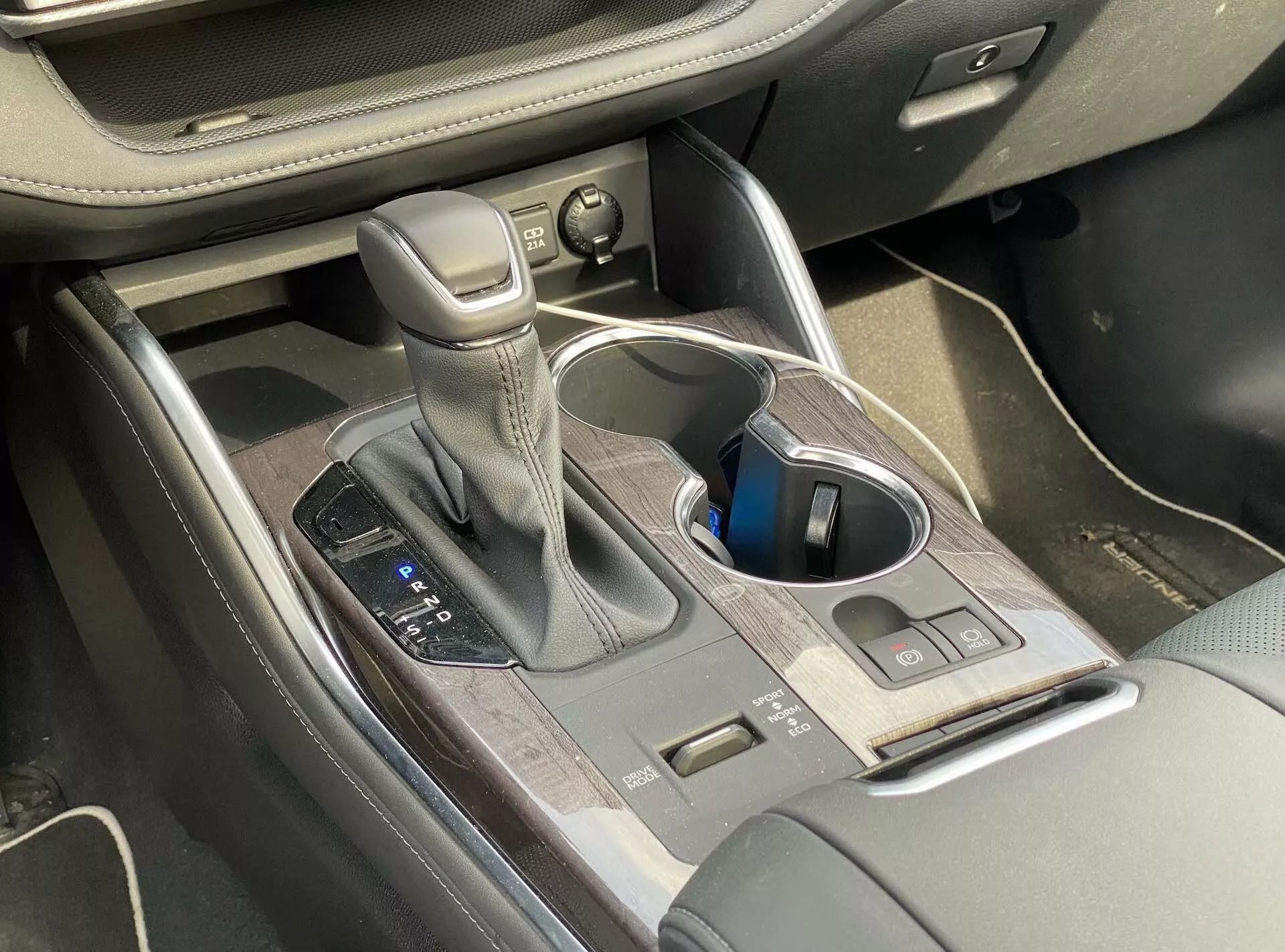 Vue intérieure du Toyota Highlander 2.5 Hybrid Business Edition 2021 focalisée sur la console centrale, montrant le levier de changement de vitesse automatique avec des coutures en cuir, les commandes de modes de conduite, les porte-gobelets, les boutons de contrôle, le rangement de la console centrale avec finition en bois, et les commandes de réglage du siège partiellement visibles.