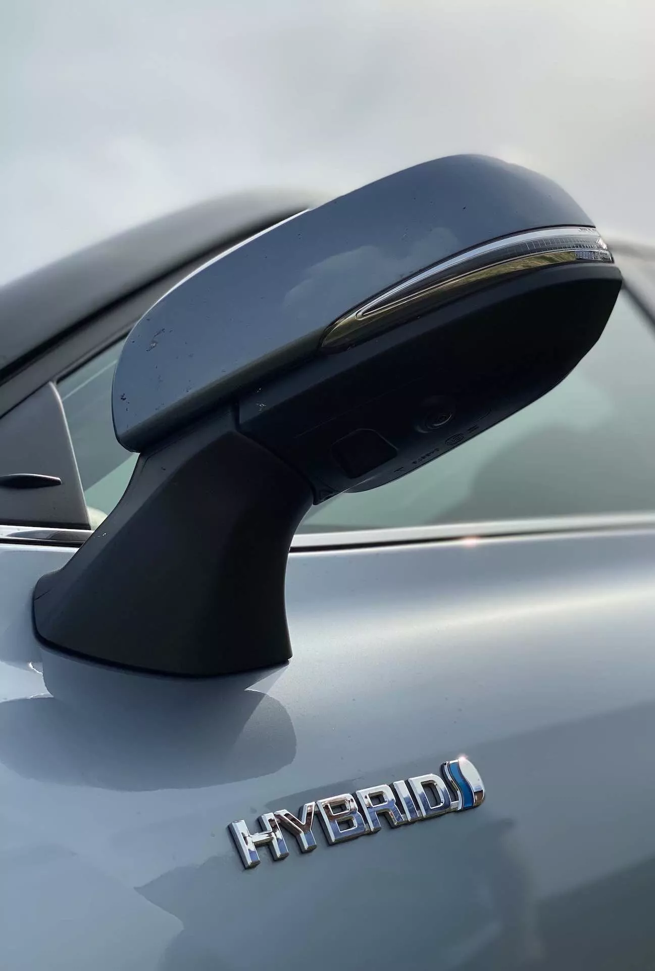 Gros plan sur le rétroviseur extérieur côté conducteur et vue partielle latérale d'un Toyota Highlander 2021 Hybrid Business Edition. Le rétroviseur, monté sur la porte, présente un design aérodynamique avec un clignotant intégré. La photographie, prise en contre-plongée, met en avant le badge 'HYBRID' en majuscules indiquant la motorisation hybride écologique. La finition métallisée grise du véhicule est clairement visible. Les détails nets sont accentués par une faible profondeur de champ qui floute l'arrière-plan.