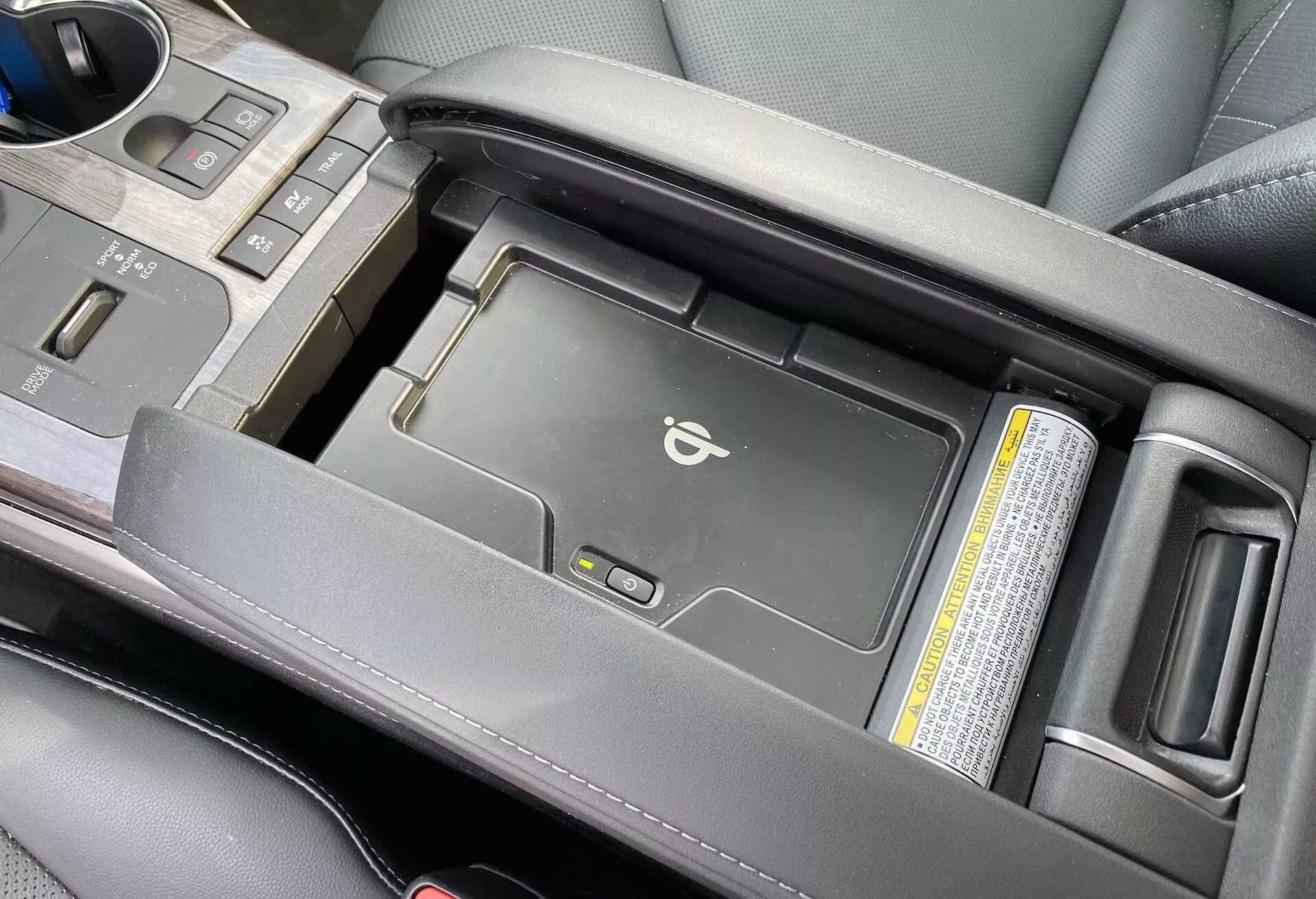 Vue rapprochée de la console centrale du Toyota Highlander 2021 Hybrid Business Edition sous un angle légèrement élevé, montrant l'aire d'accoudoir central partiellement ouverte pour révéler un chargeur sans fil pour appareils mobiles. L'image met en valeur l'accoudoir en cuir noir premium, le levier de vitesse et les porte-gobelets, en se concentrant sur les commodités technologiques modernes et la qualité esthétique.
