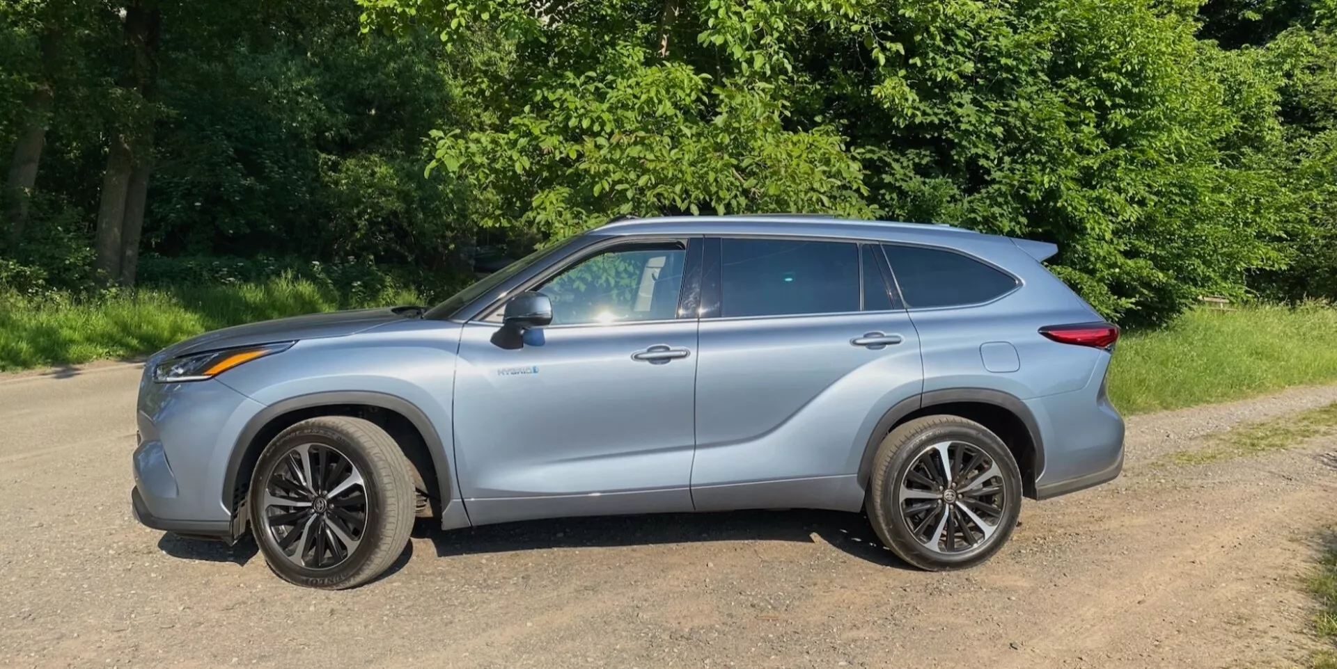 Vue de profil latéral d'un crossover SUV Toyota Highlander 2.5 Hybrid Business Edition 2021 avec une carrosserie argent métallisé, garé sur une surface en terre ou en gravier, entourée de feuillage vert dans un parc naturel ou une zone rurale. Le véhicule présente un design moderne et aérodynamique avec des lignes de contour et une forme de fenêtre profilée et élégante. Les rétroviseurs latéraux sont assortis à la couleur de la carrosserie et les roues compliquées à plusieurs rayons sont équipées de pneus à profil bas. Les barres de toit ajoutent praticité et style robuste, les poignées de porte visibles se fondent avec une ligne caractéristique qui les traverse, et le véhicule dégage un sens de mouvement vers l'avant même à l'arrêt.