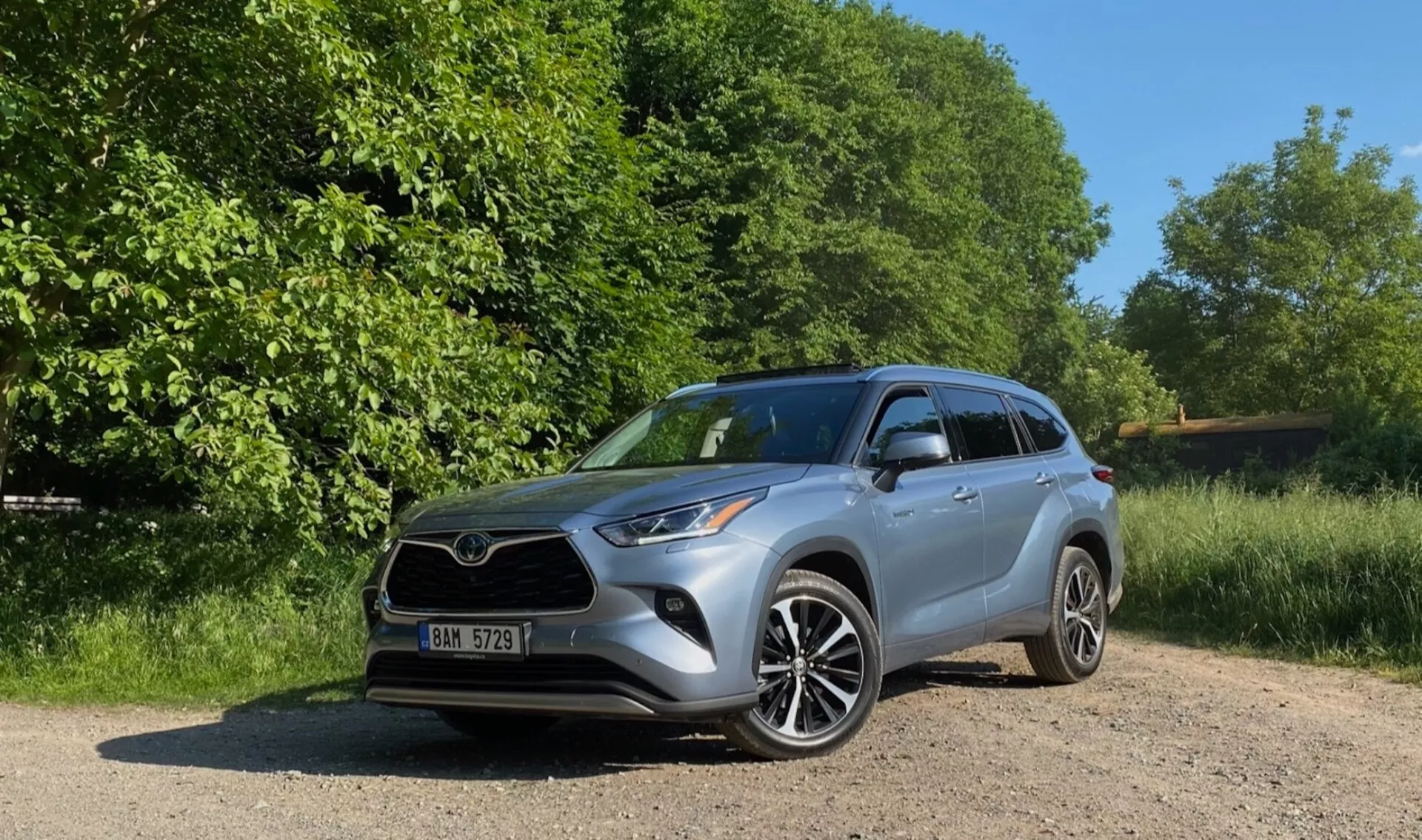 Un Toyota Highlander 2.5 Hybrid Business Edition argenté de 2021 est garé sur un chemin de gravier entouré d'arbres et de buissons luxuriants. La journée bien ensoleillée met en valeur les éléments de design détaillés du véhicule, y compris les phares à LED, les roues en alliage à rayons divisés et les clignotants intégrés dans les rétroviseurs. L'image est prise d'un angle bas en vue de trois quarts avant, en soulignant la posture robuste du SUV, la calandre avant, les phares et le profil latéral.