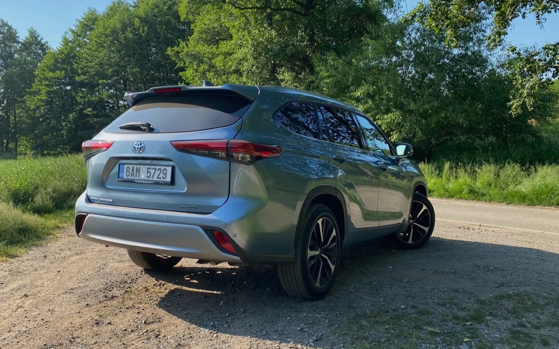 Vue arrière trois quarts d'un SUV Toyota Highlander 2021 2.5 Hybrid Business Edition, capturée depuis le niveau du sol avec un angle vers le haut. L'image met en évidence le côté arrière droit, y compris le pare-chocs arrière, le feu arrière, le hayon avec spoiler sur le toit, la porte passager, la fenêtre et la roue arrière. La peinture métallique du SUV brille au soleil, positionnée sur une surface non pavée entourée de verdure.