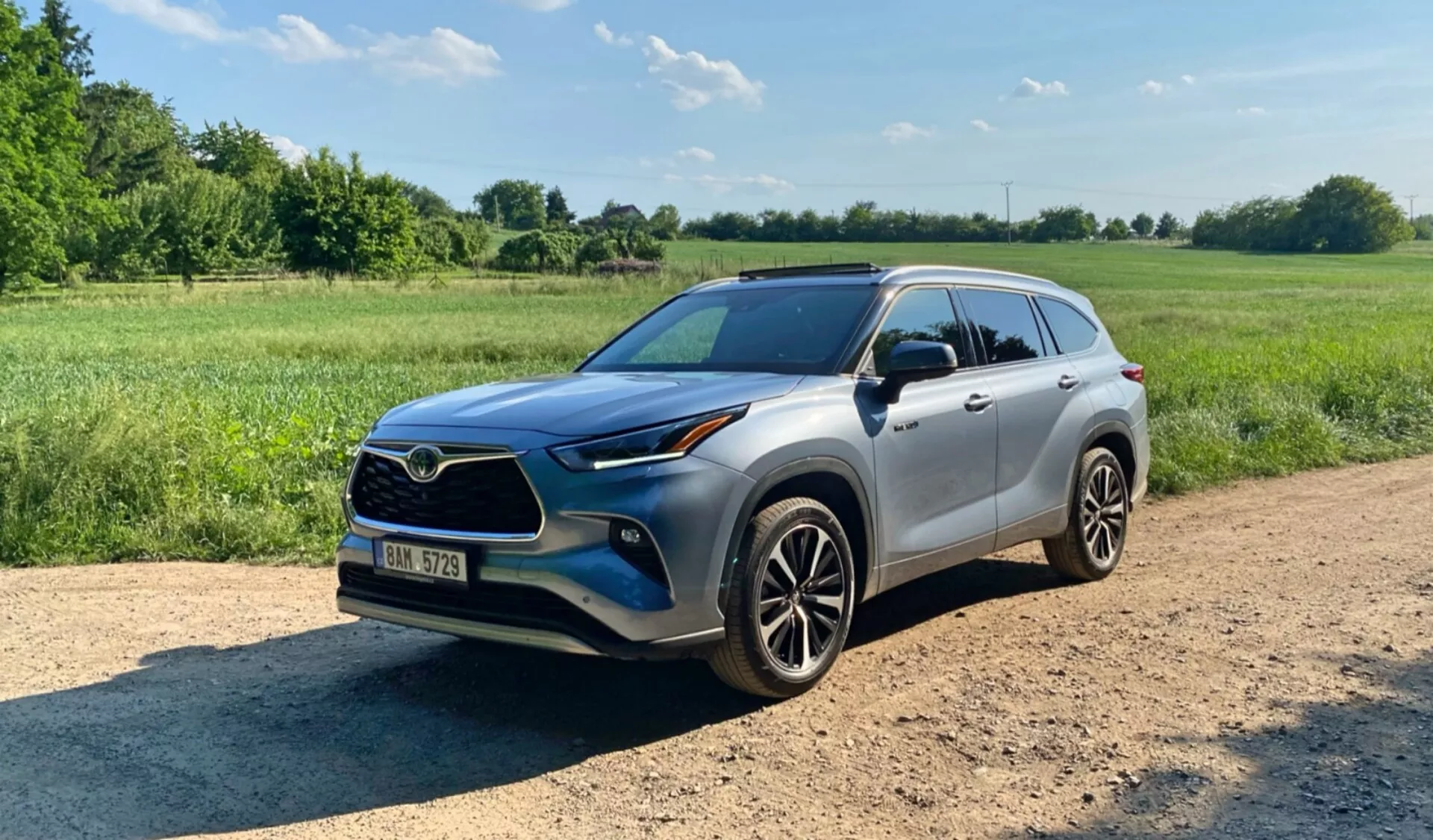 Image d'un SUV Toyota Highlander 2.5 Hybrid Business Edition 2021 prise de trois quarts avant sur un chemin de terre avec de la végétation et un ciel dégagé en arrière-plan. La photo met en évidence la calandre avant, les phares, le capot, les ailes avant, la porte côté conducteur, les rétroviseurs latéraux, les roues et les barres de toit du véhicule. Il dispose de jantes en alliage modernes, de phares LED ou de projecteur, de rétroviseurs latéraux assortis à la carrosserie avec des clignotants intégrés potentiels, d'un toit ouvrant et de vitres teintées. Le SUV est conçu avec une garde au sol plus élevée et des éléments pratiques comme des feux antibrouillard et des prises d'air sur le pare-chocs avant.