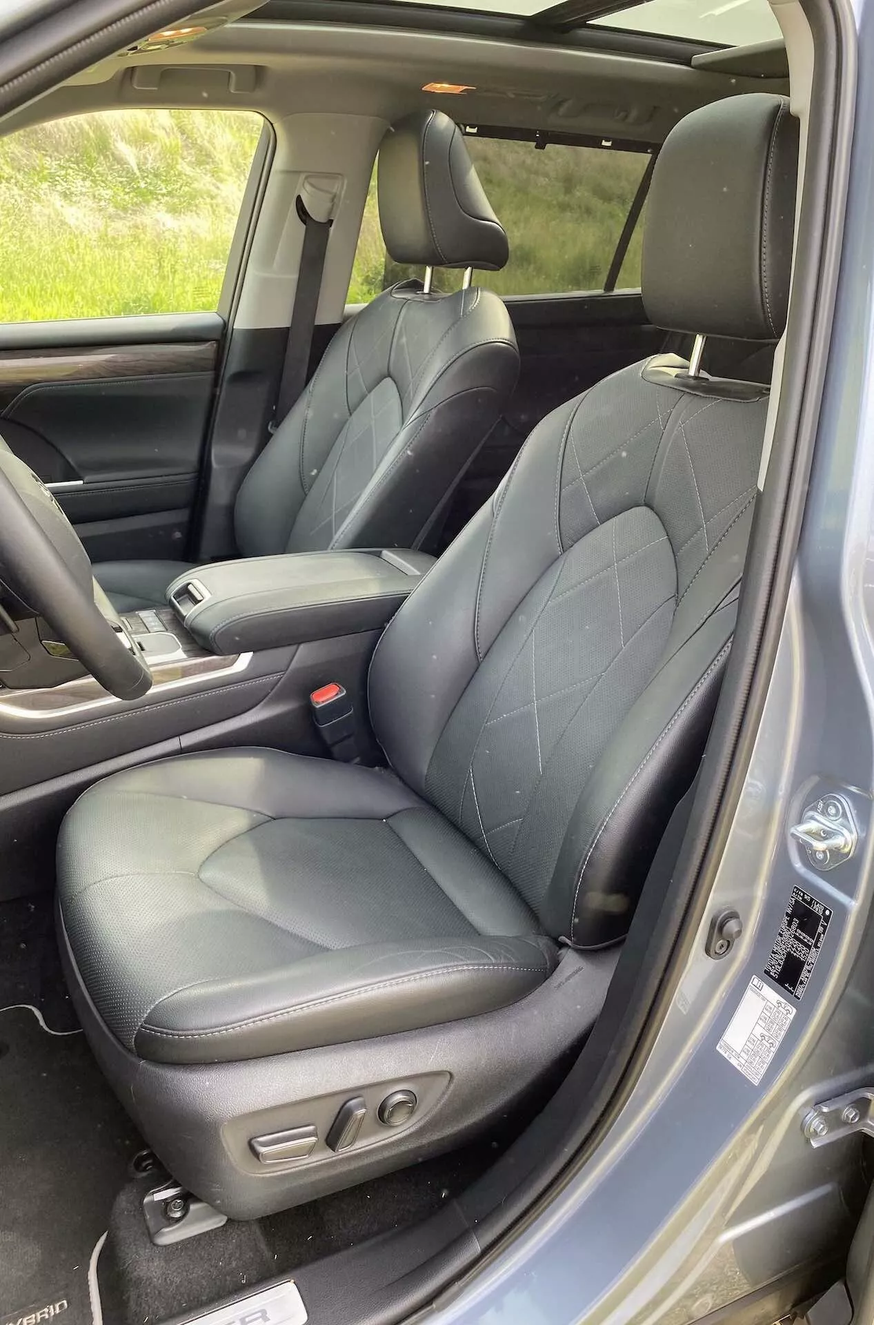 L'intérieur de la Toyota Highlander 2021 2.5 Hybrid Business Edition, montrant la zone du siège conducteur avec une sellerie en cuir noir matelassé, des renforts latéraux et des commandes de réglage électrique. La vue comprend une partie de la console centrale, le panneau de porte avec des commandes pour les vitres et rétroviseurs électriques, la grille de haut-parleur et la ceinture de sécurité avant. Le design luxueux et ergonomique est mis en valeur par la lumière naturelle.