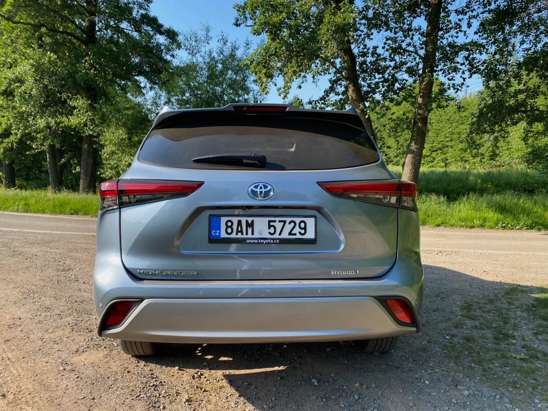 Vue arrière d'une Toyota Highlander 2.5 Hybrid Business Edition argentée affichant les badges HYBRID et AWD-i, la plaque d'immatriculation centrale, l'emblème Toyota et les feux arrière avec un insert réfléchissant, le tout sur une surface en asphalte avec un feuillage vert en arrière-plan.