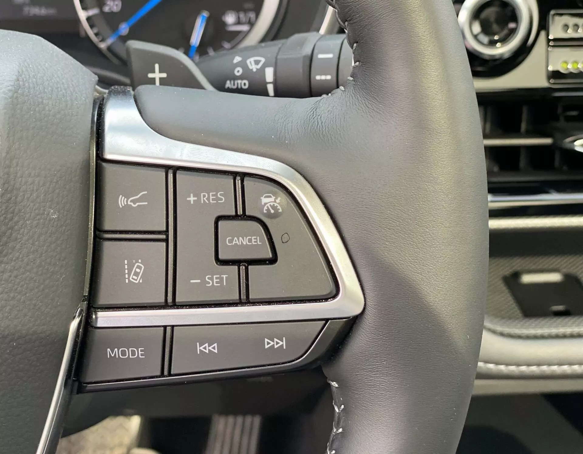 Gros plan sur le côté gauche du volant d'une Toyota Highlander 2021 2.5 Hybrid Business Edition, présentant du cuir noir avec des coutures visibles, des accents en argent/chrome, des boutons de commande de régulateur de vitesse adaptatif et du système multimédia, et une partie du tableau de bord et du panneau central.