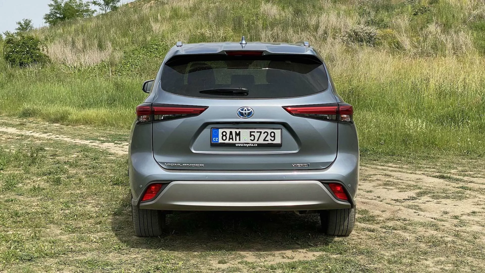 Vue arrière d'une Toyota Highlander 2021 2.5 Hybrid Business Edition de couleur argentée, garée sur une zone herbeuse avec un sentier poussiéreux au premier plan, capturée à la lumière du jour avec un éclairage naturel. Le SUV moderne présente des feux arrière élégants enveloppant les côtés, un spoiler arrière proéminent, des réflecteurs intégrés dans le pare-chocs, une caméra de recul au-dessus de la plaque d'immatriculation et une plaque de protection argentée sous le pare-chocs, suggérant ses capacités tout-terrain et les avancées technologiques en matière d'assistance au stationnement et de systèmes de recul.