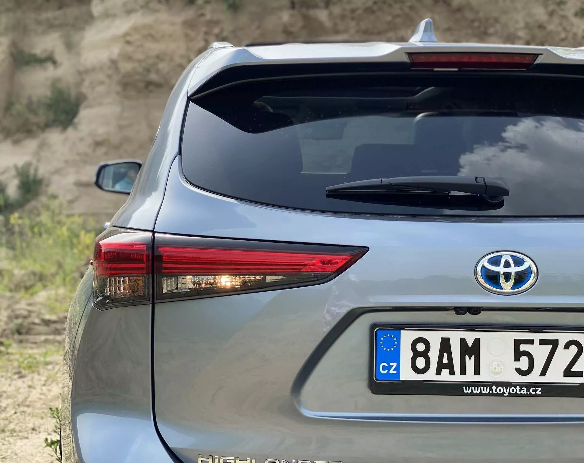 Gros plan de la section arrière du Toyota Highlander 2021 Hybrid Business Edition en argent, capturé sous un angle de trois quarts mettant en valeur le côté droit. Il présente un feu arrière à LED distinctif, une plaque d'immatriculation de style européen de République tchèque, un badge hybride, un système de dégivrage de la lunette arrière et un troisième feu stop monté en hauteur contre un arrière-plan de terrain naturel.