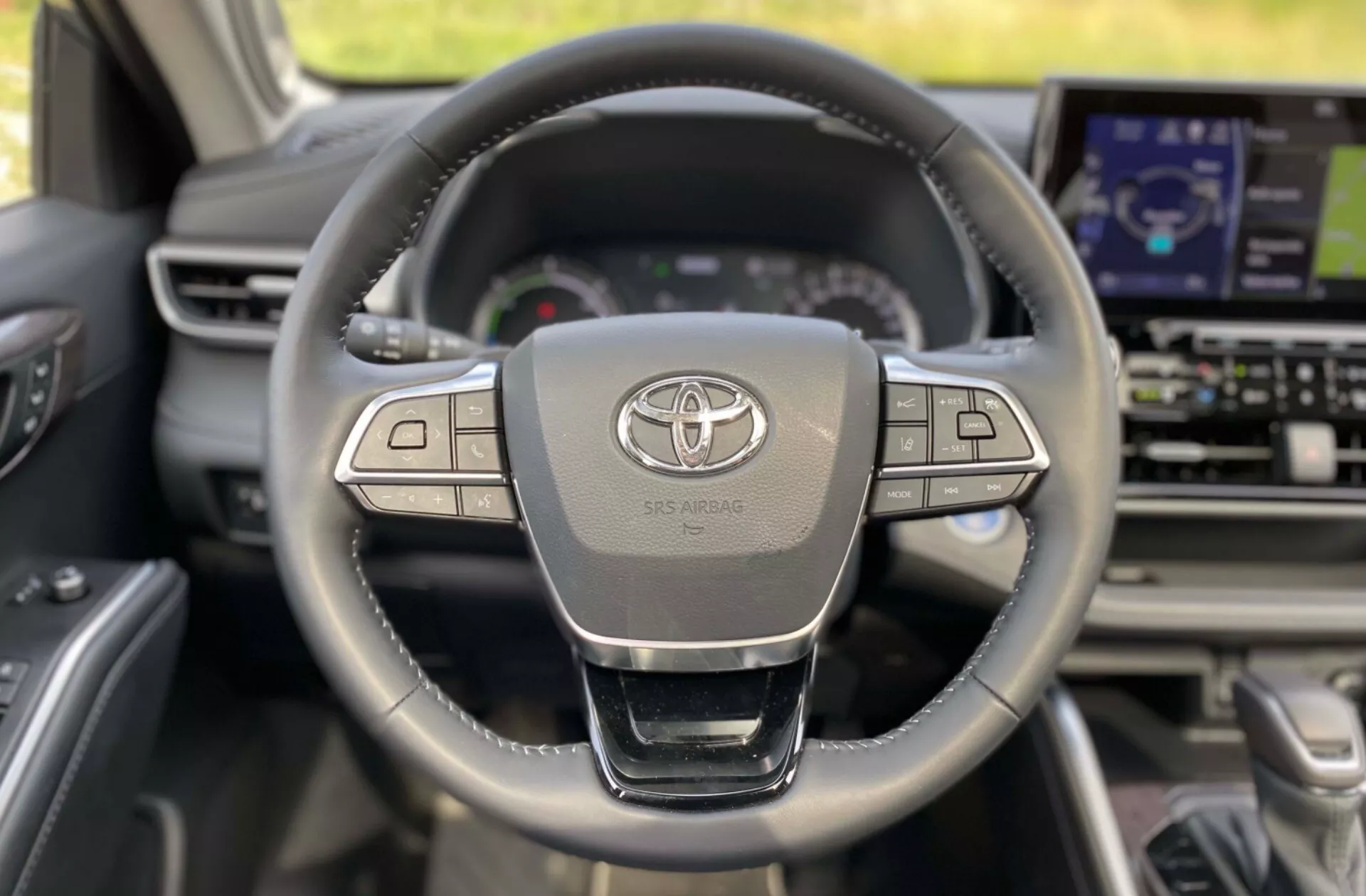 Intérieur d'une Toyota Highlander 2021 Business Edition vue de la perspective du conducteur, avec un volant en cuir à trois branches avec plusieurs boutons de contrôle, le logo Toyota et l'inscription SRS AIRBAG. En arrière-plan, le tableau de bord comprend un afficheur numérique et un écran d'infodivertissement plus grand.
