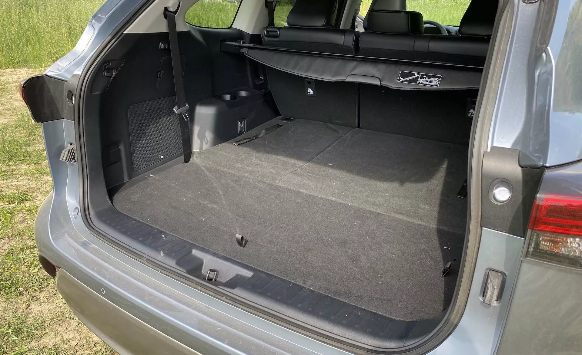 Vue rapprochée de la zone de chargement ouverte du Toyota Highlander 2.5 Hybrid Business Edition 2021 avec hayon vertical et compartiment arrière spacieux. L'angle depuis le côté passager montre une surface propre et moquettée, les sièges arrière pouvant être abaissés pour agrandir l'espace de chargement. Les caractéristiques visibles incluent une lumière intérieure, une prise de courant 12V, une languette pour accéder à un espace de rangement supplémentaire, des ancrages pour sièges enfant et une partie du groupe de feux arrière à LED modernes.