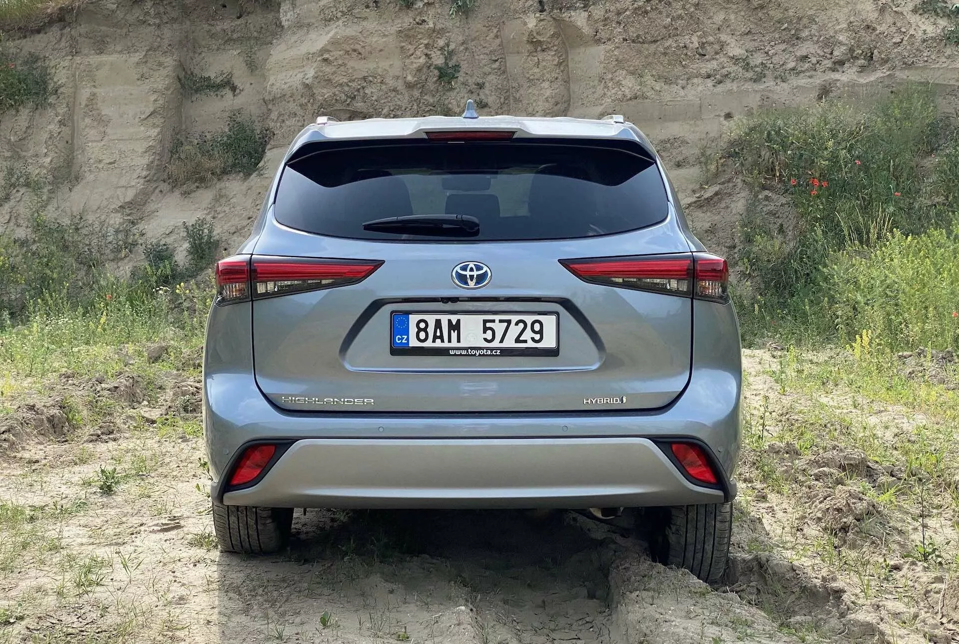 Vue arrière d'une Toyota Highlander 2021 2.5 Hybrid Business Edition argentée capturée à hauteur des yeux, montrant le hayon avec l'emblème Toyota, les feux arrière, système d'échappement double, plaque d'immatriculation au centre du pare-chocs arrière et le badge hybride, le tout sur un terrain naturel avec de l'herbe et de la terre, soulignant son potentiel tout-terrain.